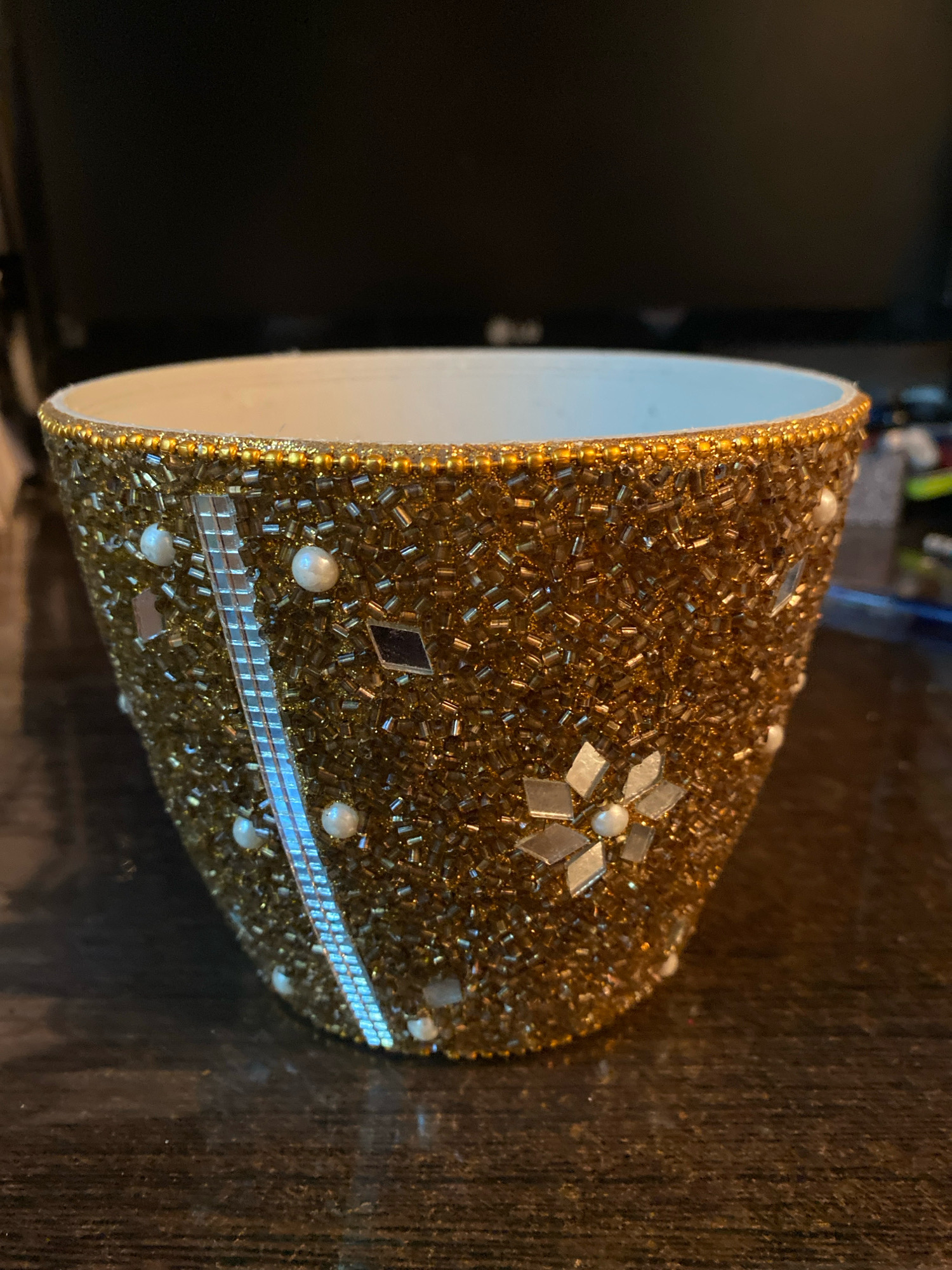 A close-up, eye-level shot shows a decorative gold-beaded pot on a dark wood surface. The pot has a white interior and is adorned with pearls and mirrored embellishments. A thin band of gold beads encircles the rim. The background is dark and out of focus, with hints of electronic equipment visible. The image was captured in Ivanteyevka, Russia.