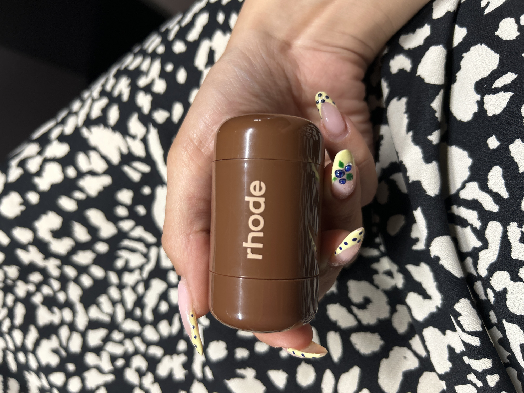 A person is holding a brown, cylindrical object with the word "rhode" printed in white letters on its side. The object is oval-shaped and appears to be a cosmetic product. The person's hand has light skin and is decorated with a French manicure with yellow tips and blue dots, and some nails have blueberry designs. The person is wearing a black and white patterned garment that is draped around them. The scene is an indoor setting, possibly a home or studio, with no discernible landmarks indicating a specific location in Tbilisi, Georgia.