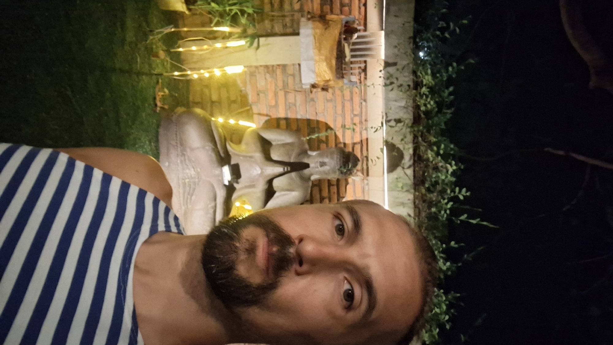 A man with a beard and a striped tank top is taking a selfie at night. In the background, there is a Buddha statue and some decorative lights. The location is unknown.