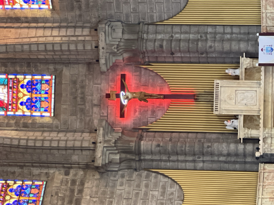The image depicts the interior of Nha Trang Cathedral (also known as the Stone Church) in Nha Trang, Vietnam, specifically focusing on the main altar area. Dominating the center is a large crucifix featuring a sculpted figure of Christ, illuminated by a prominent red light emanating from behind the cross and a glowing white halo around Christ's head. Below the crucifix, a stone altar structure is visible, flanked by two small white statues, possibly angels, each appearing to hold a light source or votive candle. Behind and to the sides of the altar and crucifix, numerous vertical golden pipes, characteristic of a large pipe organ, are arranged in a semi-circular or curved pattern. The surrounding walls are constructed from large, rough-hewn grey stone blocks, exhibiting visible mortar joints and architectural details such as fluted columns and decorative cornices. Above the altar, two large stained-glass windows are visible, depicting religious figures in vibrant blues, reds, and yellows, with text in French reading "LE CHRIST ROI" (Christ the King) on the right window. A small portion of a white banner with a coat of arms or religious emblem is visible at the bottom right of the frame. No human individuals are visible. The scene is static and presents a solemn religious setting.