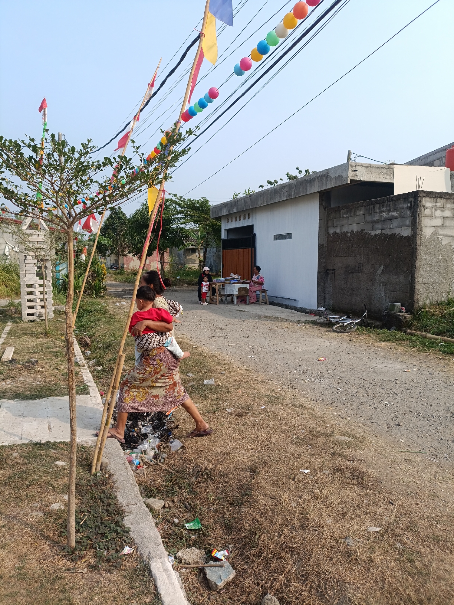 The image depicts a street scene in Cariu, Indonesia. A woman, possibly of adult age, is seen carrying a young child on her back. She is wearing a patterned skirt and a red shirt. The child is also wearing a red shirt. They are standing near a small tree adorned with colorful decorations and flags. In the background, there are residential buildings, one with a white facade and another constructed with cinder blocks. A few other individuals are visible in the distance, some seated at a table outside a building. A bicycle is lying on the ground near the buildings. The environment appears to be a rural or semi-urban area, with a dirt road and dry grass. There is some debris scattered on the ground, including plastic waste. The sky is clear and blue.