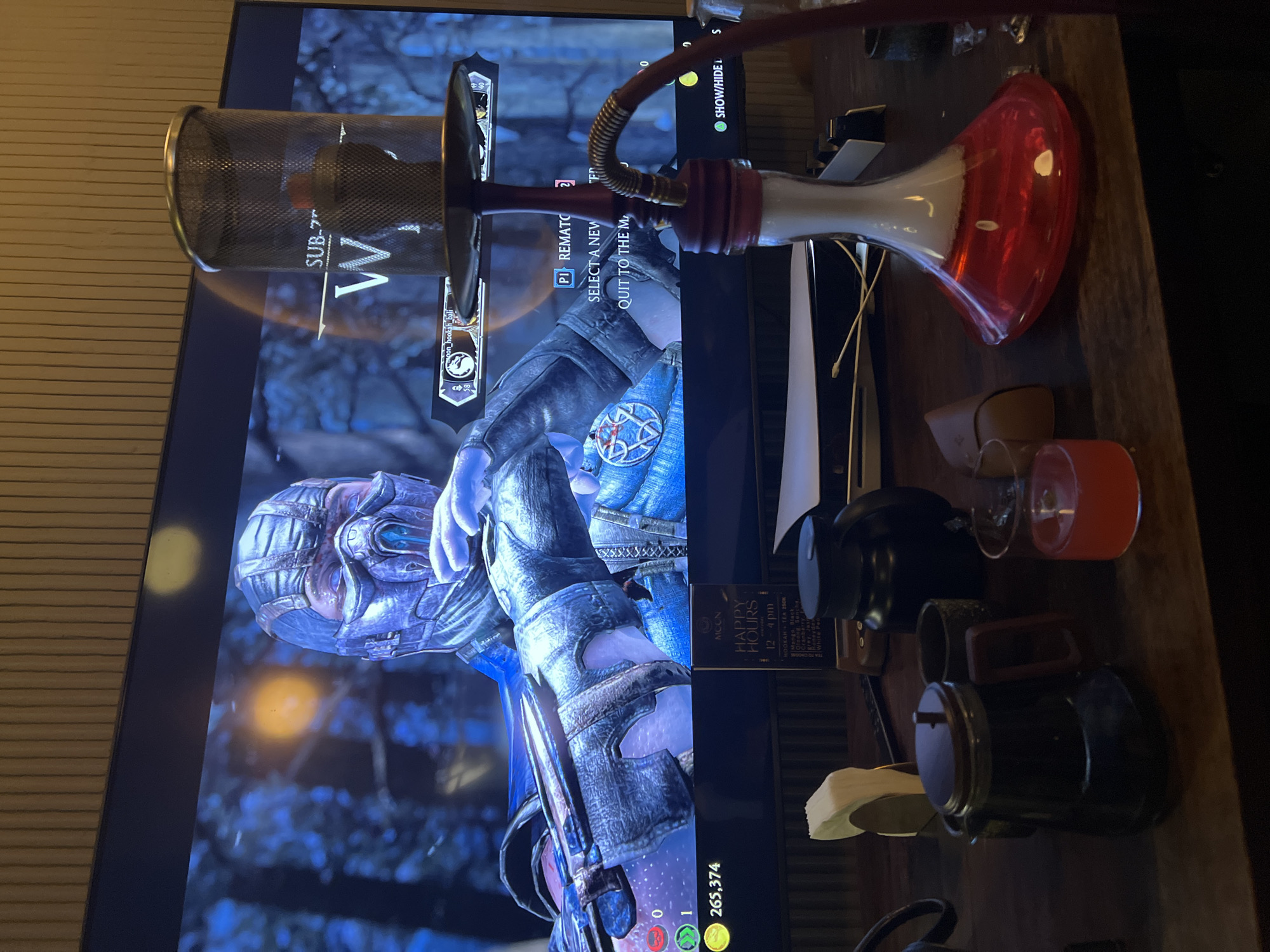 The image shows a television screen displaying the character Sub-Zero from the Mortal Kombat game. In front of the screen, on a table, is a hookah with a red and white base. Next to the hookah are various items, including glasses, a black device, and a card that reads "MCON HAPPY HOURS, 12-4 pm, HOOKAH TEA 250K". The card also has "noon_hookah bali" written on it, suggesting that the photo was taken at or near this location in North Kuta, Indonesia. A game menu is displayed on the television screen.