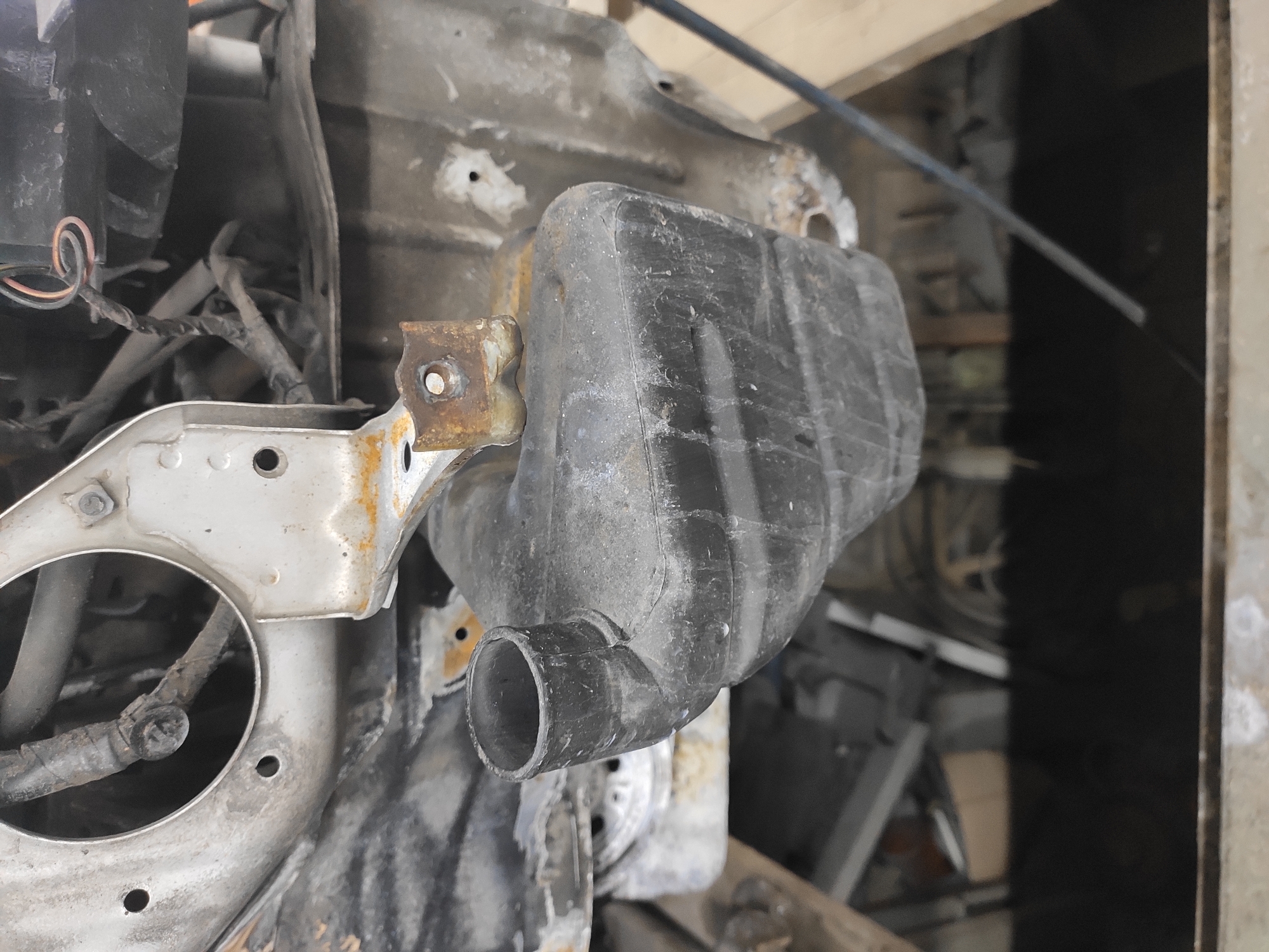 The image shows a close-up, low-angle view of automotive components, likely from the front of a vehicle. A black plastic reservoir with a circular opening at the top is prominently featured, suspended by a rusty metal bracket. Behind it, various metallic structures, wires, and hoses are visible, suggesting an engine bay or undercarriage. The overall impression is one of disrepair and partial disassembly, with evident signs of rust and dirt. The context of Homyel, Belarus is not discernible from the visual information provided in the image.