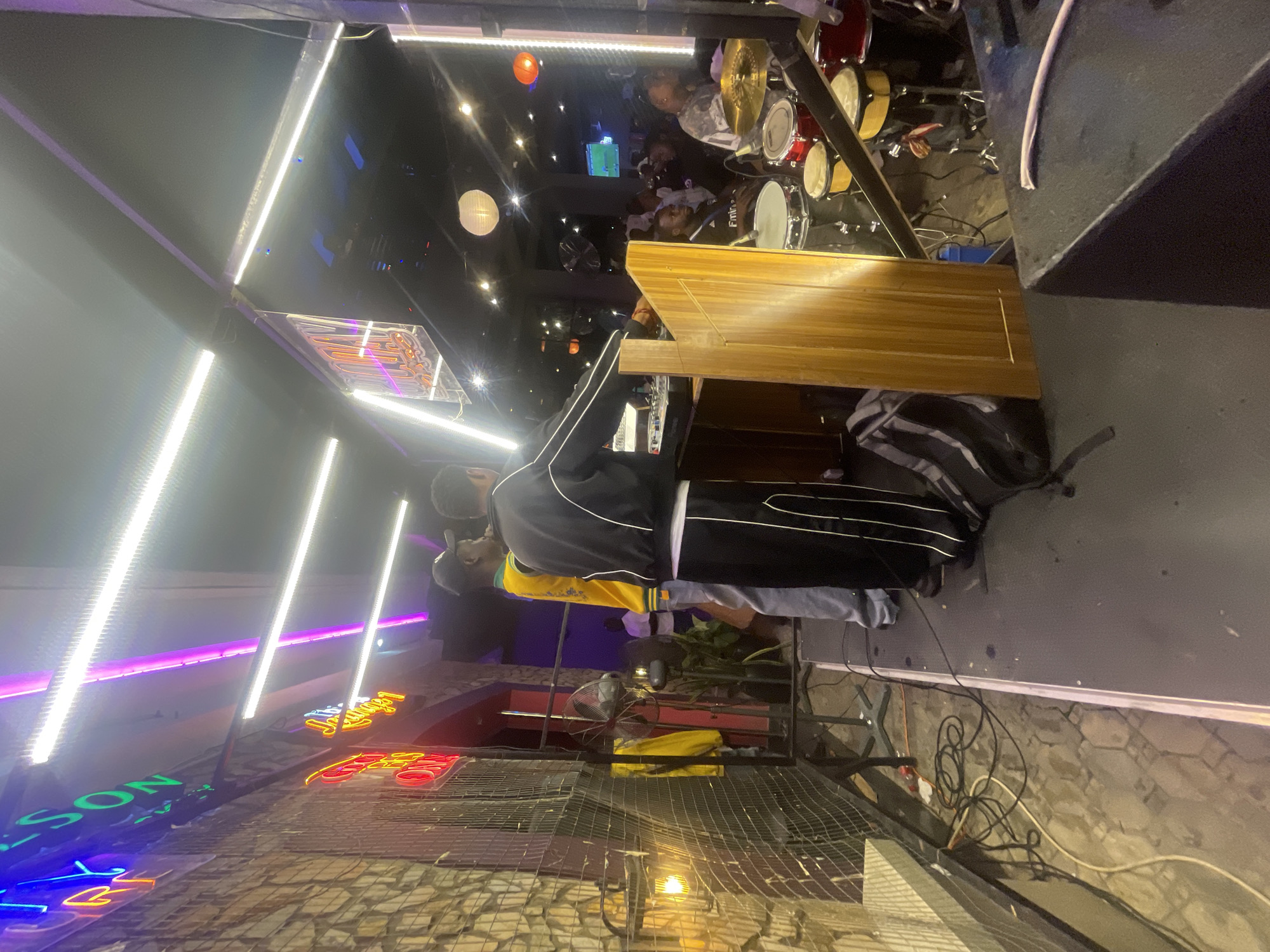 The image shows the inside of a bar or nightclub, potentially in Lokoja, Nigeria, given the presence of African individuals and a humid, tropical environment. A DJ is operating a sound mixing console. Behind him are the drums of a band. There are people sitting around the interior of the establishment. Bright, colorful neon signs and LED lights illuminate the scene. The flooring appears to be a combination of tile and concrete. A metal fence in the foreground separates the space from an outside area.