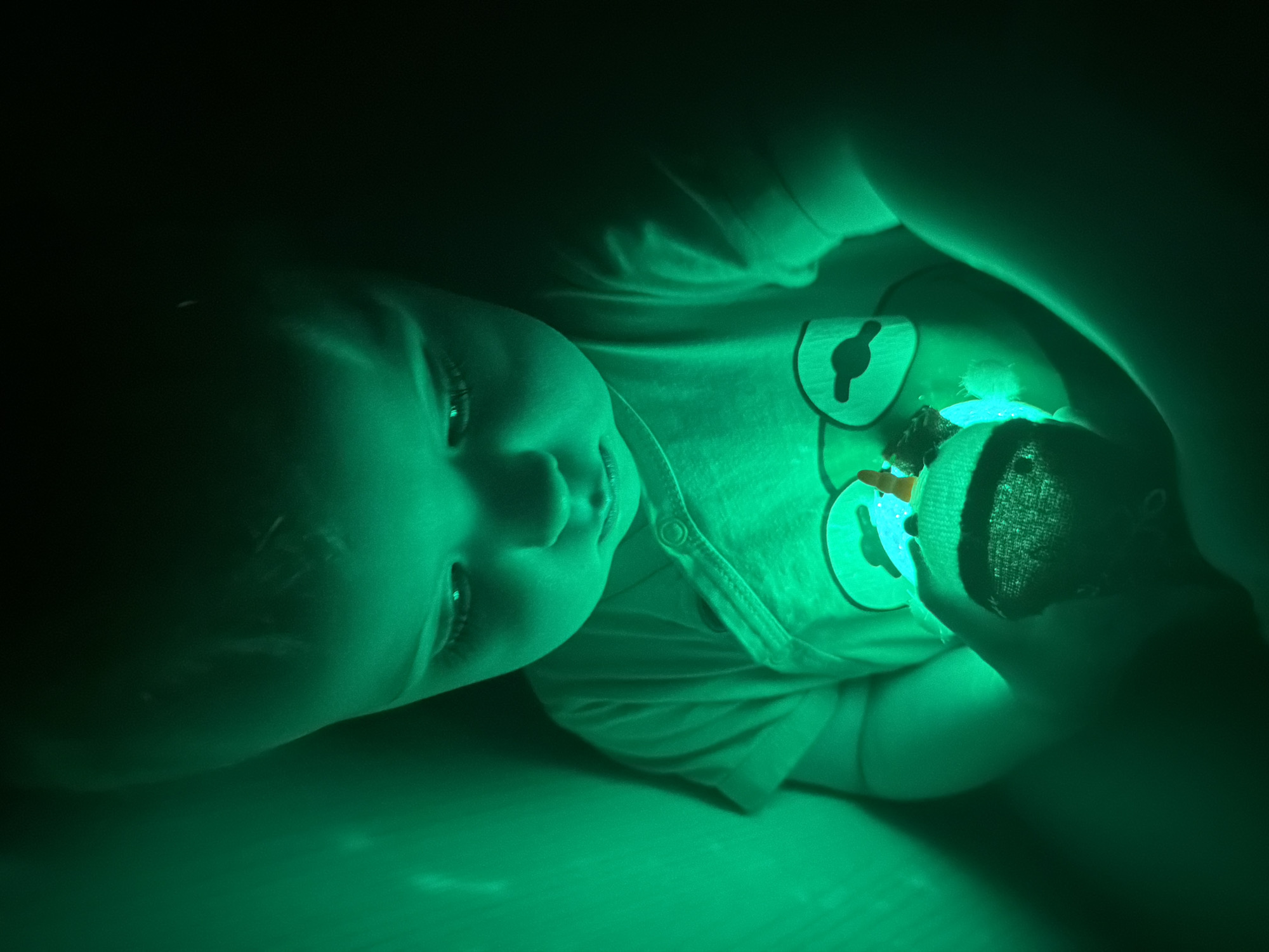 A child is lying on their back in near darkness, with their face angled towards the left and lit by a soft green glow. The child is wearing a light-colored shirt with button details. The shirt features a design of animal shapes. The child is holding a toy in their hands, which is the source of the green light. The environment surrounding the child is dark, creating a sense of confinement and focused attention on the illuminated face and toy. The location of the image in the city of Votkinsk, Russia cannot be determined.