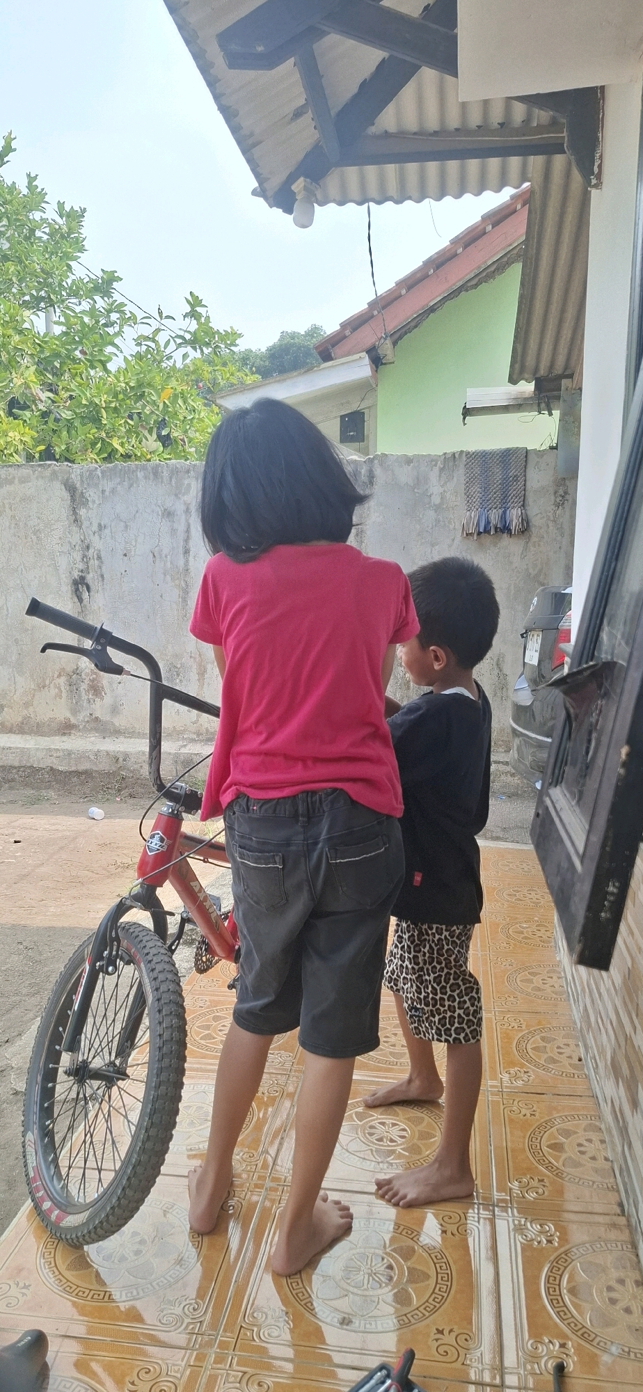 In the city of Cibinong Hilir, Indonesia, two children are standing on a tiled patio next to a red BMX bicycle. The child on the left, appearing to be a girl with short black hair, is wearing a pink t-shirt and dark gray shorts. She is facing away from the camera, with her left hand on the handlebars of the bicycle. The child on the right, appearing to be a boy, is wearing a black t-shirt and patterned shorts. He is also facing away from the camera and stands slightly behind the first child. The bicycle is red with black tires and handlebars. In the background, there is a concrete wall, a green building with a red-tiled roof, and lush green foliage. The sky is bright and appears to be daytime.