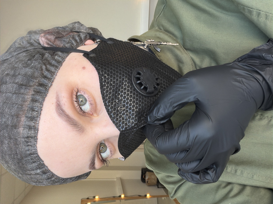 A person wearing a gray hairnet and black face mask with a round vent is seen in a close-up shot. They wear black gloves and an olive-green shirt or jacket adorned with a silver gun pin. The person's face, particularly their eyes and eyebrows, are in focus. The background is indistinct, showing only a blurred glimpse of a light-colored wall and a row of small lights.