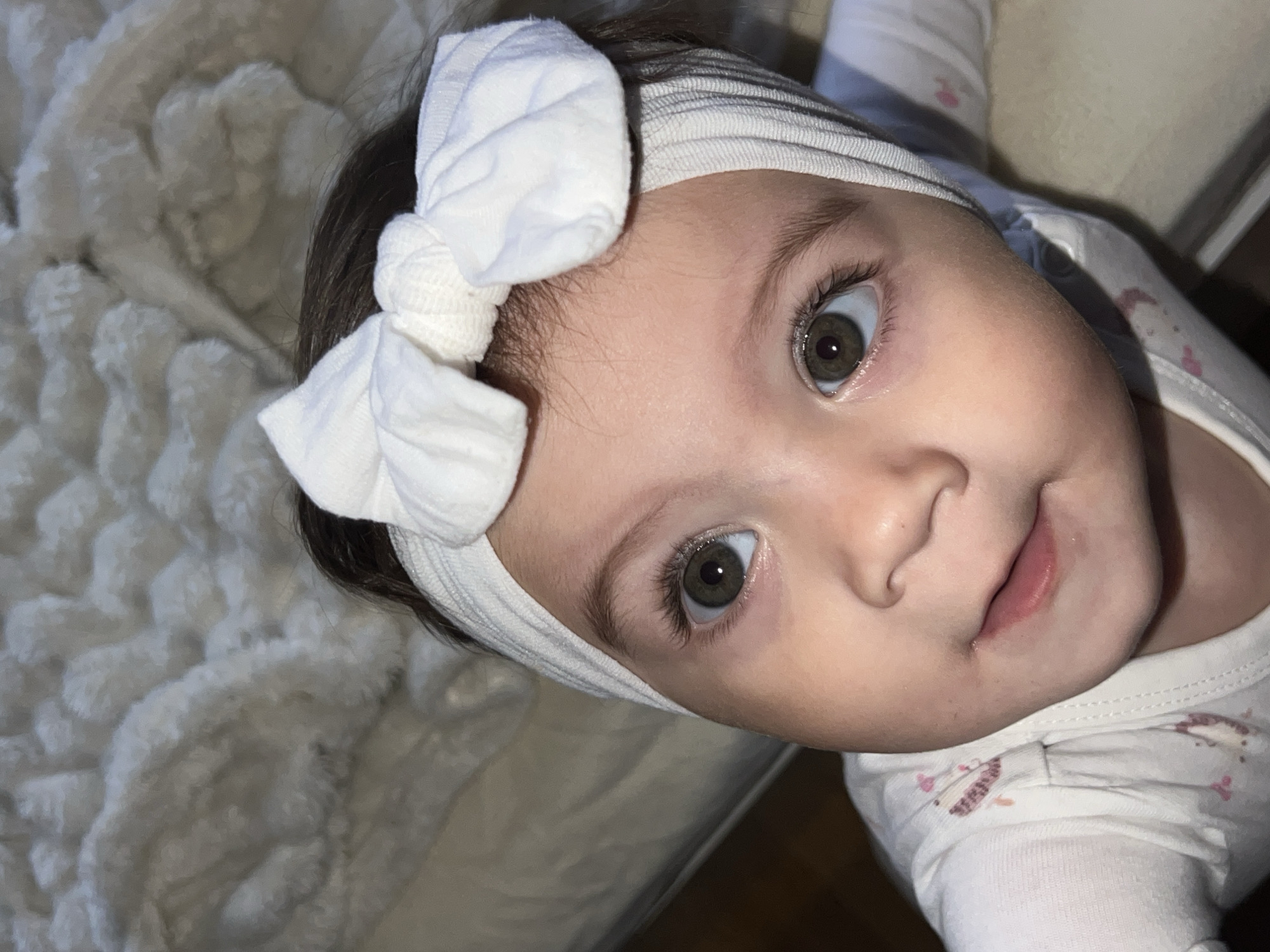 A high-angle, close-up photograph depicts an infant, approximately six to eighteen months of age, situated in an indoor environment in the city of Elena, Bulgaria. The subject has dark hair, prominent dark green or hazel eyes, light skin, and is looking directly upwards towards the camera with a neutral expression and slightly parted lips. The infant wears a white ribbed headband adorned with a large white fabric bow positioned on the crown of the head. Clothing consists of a white long-sleeved garment, possibly a onesie, featuring faint patterns of pink and brown shapes on the visible left shoulder and chest area. The immediate background is composed of a light-colored, textured, soft fabric or blanket with a raised, fluffy pattern, suggesting a bed or soft surface. A flatter, lighter surface, consistent with bedding, is visible to the upper left. The lighting is soft and ambient. No other individuals or specific objects are identifiable within the frame.