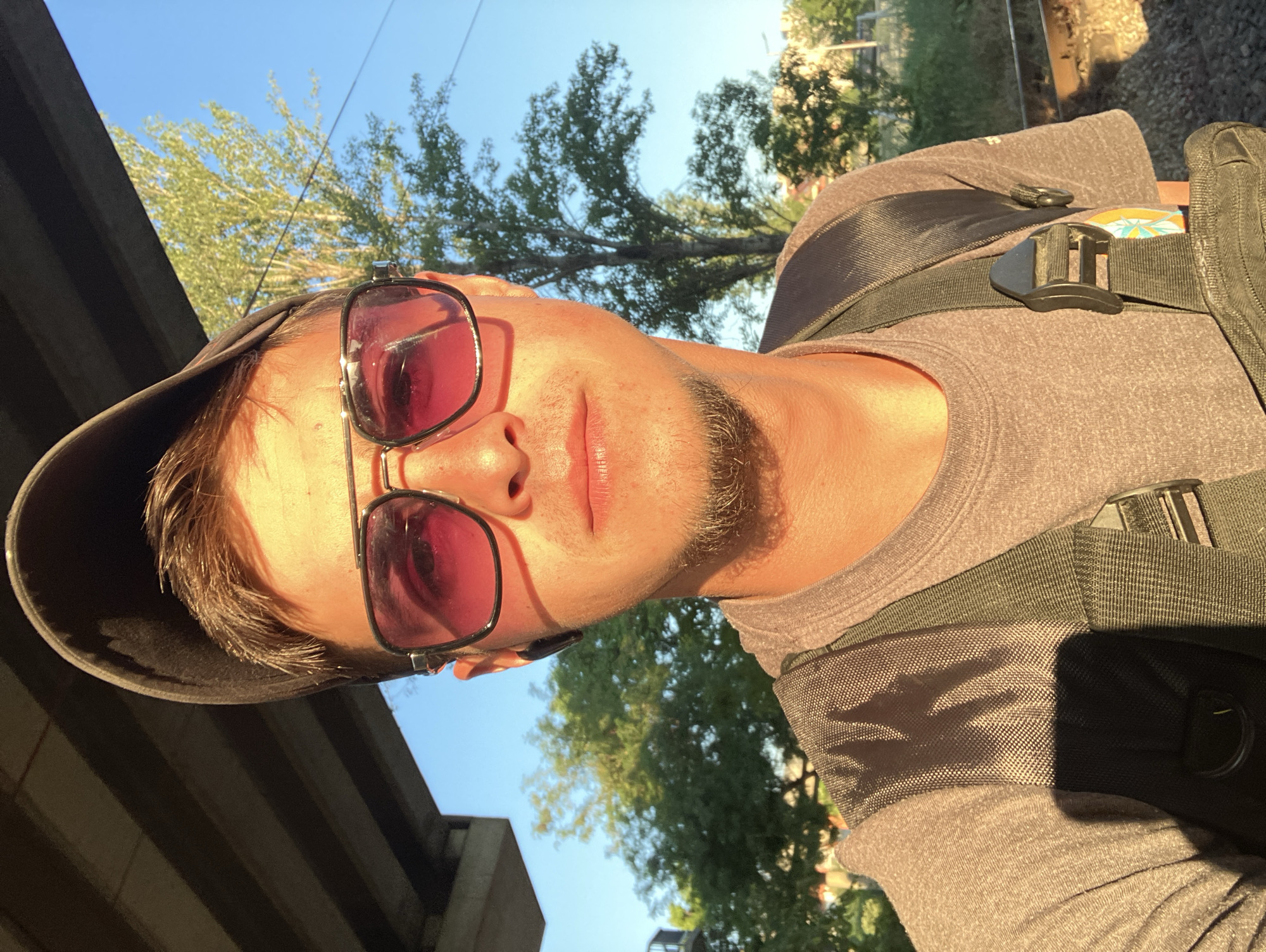 A selfie of a man wearing sunglasses and a baseball cap, situated underneath a bridge, likely near train tracks given the partial view in the upper right corner. He wears a gray shirt with a backpack. The background includes trees and a blue sky. Based on the information provided, the location is likely in Plovdiv, Bulgaria, although specific landmarks are not identifiable in the photograph.