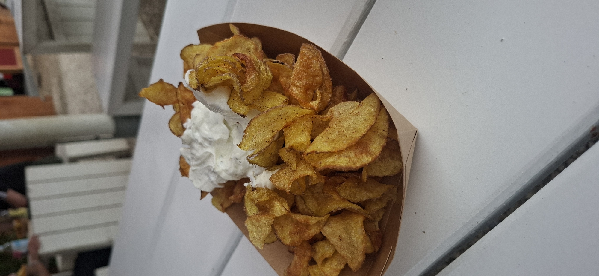 A close-up, eye-level shot shows a serving of thin, crispy potato chips with a dollop of white dip in a brown cardboard boat. The chips are golden brown and appear to be homemade. The dip is a creamy white color, possibly sour cream or yogurt-based, with visible herbs mixed in. The boat is placed on a white wooden table, with a sliver of the table’s edge visible in the foreground and background. In the blurred background, white wooden benches and a partial view of people are discernible, suggesting an outdoor seating area. The setting is likely a beer garden or outdoor restaurant. The image was taken in Hürth, Germany.