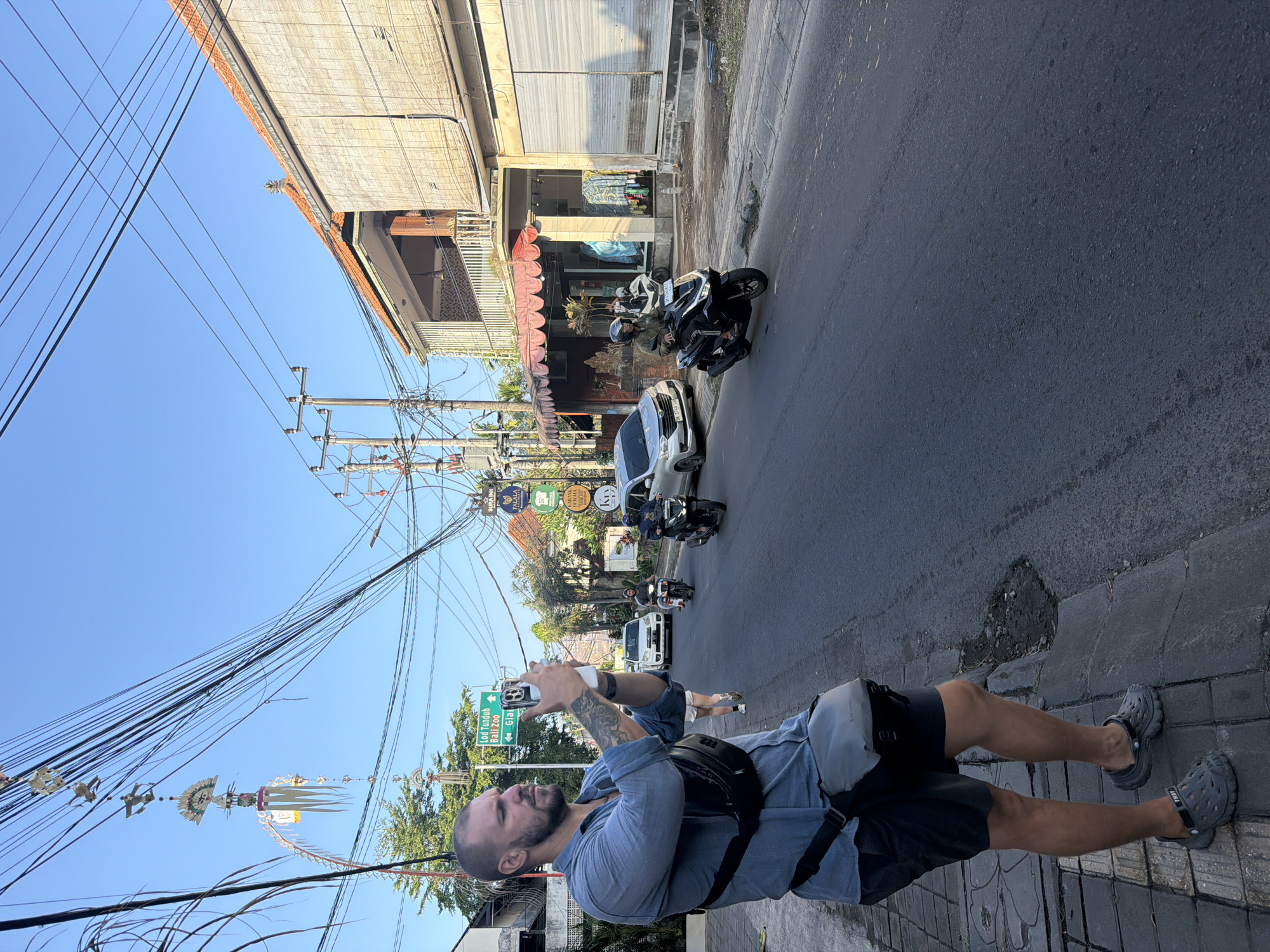 A man stands on a sidewalk, holding a smartphone up to take a picture of a street scene in Ubud District, Indonesia. He is wearing a blue collared shirt, dark shorts, a black crossbody bag, and gray Crocs. He has tattoos on his left arm. In the background, several motorbikes and cars are on the street. Buildings line the street, some with red tile roofs and bamboo blinds. Overhead, a complex network of electrical wires is visible against a clear blue sky. Traditional Balinese decorations hang from some of the wires. A green street sign indicates directions to "Loh Tunduh Bali Zoo" and "Gia".