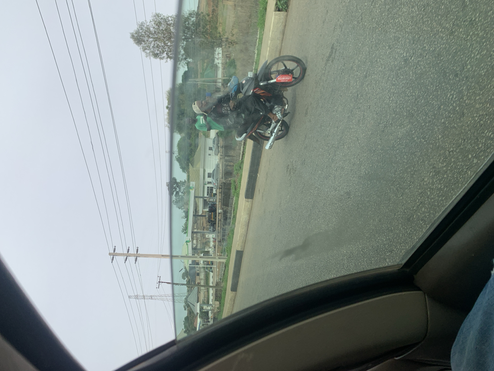 From inside a vehicle, the photo shows a motorcycle carrying two individuals moving on a road in Gura Top, Nigeria. The motorcycle is black and red. The driver is wearing black and a white cap, while the passenger is wearing green. A gas station and power lines are visible in the background. The environment appears overcast.