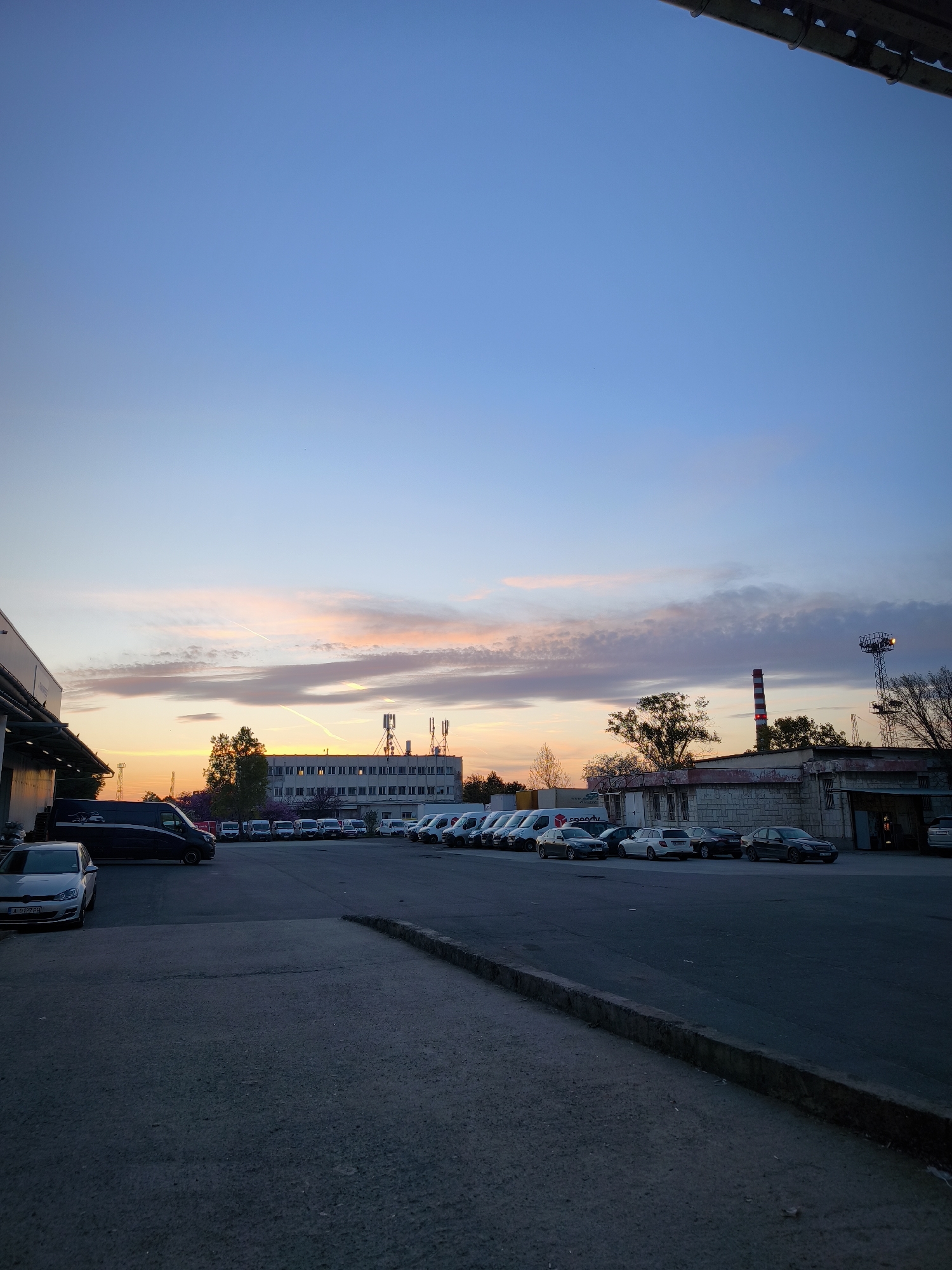The image captures an industrial or commercial parking lot in Burgas, Bulgaria, at either sunrise or sunset. The sky displays a beautiful gradient, transitioning from deep blue at the zenith to soft pinks, oranges, and yellows near the horizon, with scattered clouds and faint contrails. The overall lighting is dim, casting the foreground and buildings in shadow, enhancing the warm hues of the sky.
In the background, a multi-story building with numerous windows, possibly an office or factory, stands on the left, adorned with several cellular communication towers on its roof. To its right, a single-story building with a worn facade is visible, beside a distinctive red and white striped chimney/smokestack. Further right, a tall metal tower with lights adds to the industrial feel. Trees are interspersed between the structures.
The foreground consists of an asphalt parking area, with a raised concrete curb diagonally traversing the lower right section. Numerous vehicles are parked throughout the lot. On the left, a silver car with a Bulgarian license plate "A 0127 PH" (confirming Burgas as the location) is prominently featured, next to a dark-colored van. Further back, a line of white delivery vans is visible, one bearing the "Speedy" logo and website "www.speedy.bg", a common courier service in Bulgaria. Other parked cars, including several sedans, fill the remaining spaces. No people are visible, and no specific activity is taking place; the scene appears calm and static, reflecting the quiet hours of dawn or dusk.