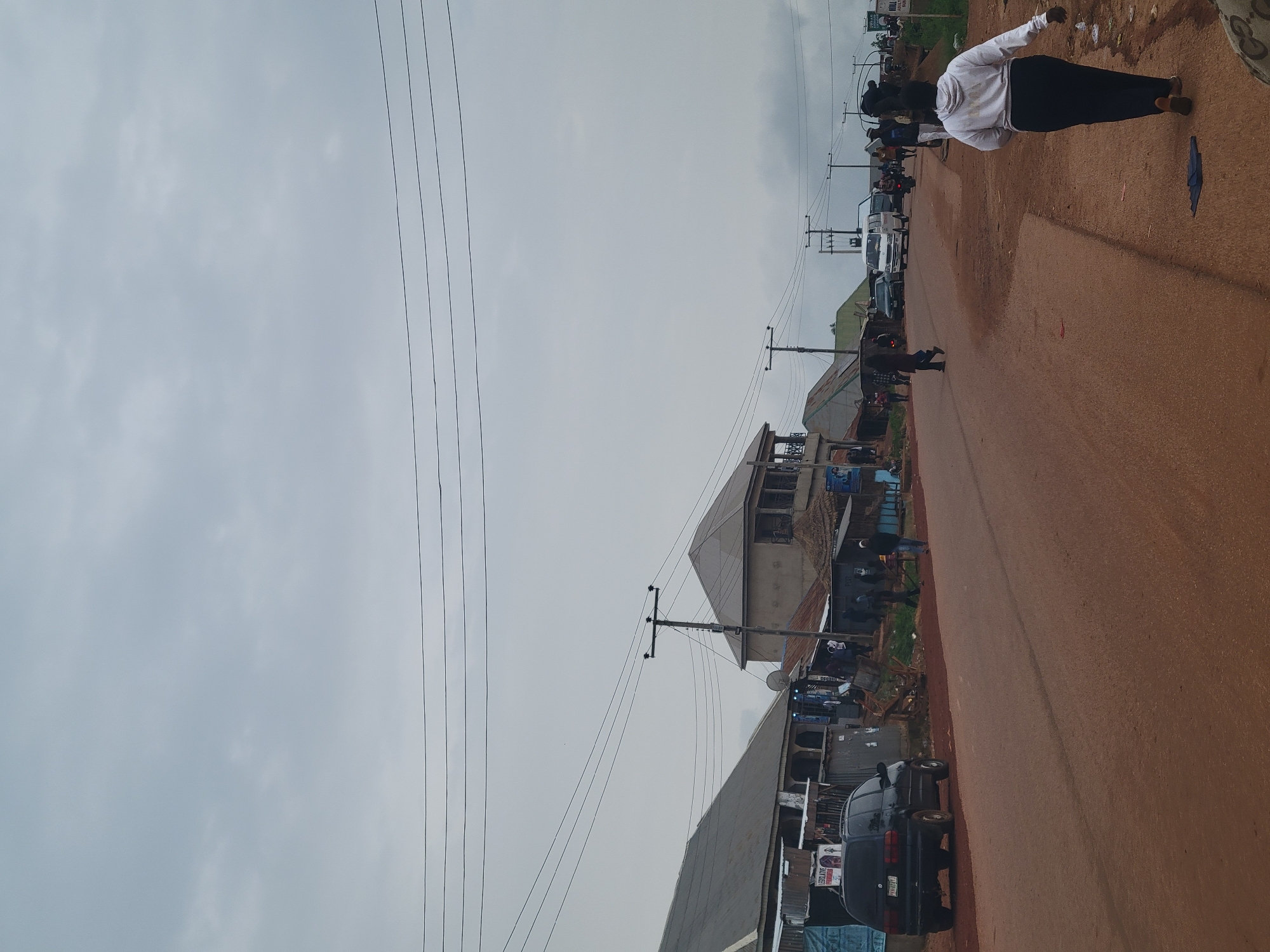 In a dusty, overcast urban environment, a street scene unfolds. The road, a light reddish-brown, is paved and appears wet in some areas. On the left side of the road, a row of modest buildings with corrugated metal roofs line the street. A black SUV is parked parallel to the curb in front of one of these structures. Further down the street, more buildings, some with balconies and thatched roofs, are visible, interspersed with utility poles and overhead wires.
Several individuals are present in the scene. In the foreground on the right, a person wearing a white hooded sweatshirt and dark pants walks away from the camera. Other people are scattered along the street, some walking, some standing near the buildings. Several vehicles are on the road, including a white van, a blue car, and a motorcycle. The sky is a dull gray, suggesting cloudy or overcast weather. The overall impression is one of a busy, everyday scene in a developing urban area.
