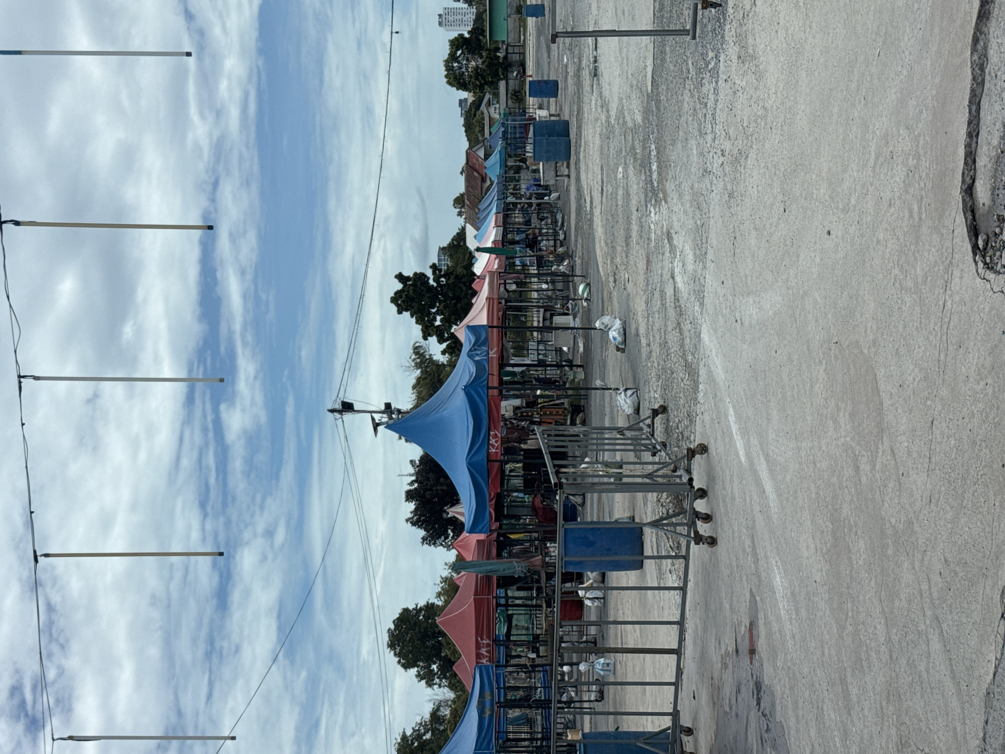 An outdoor market scene is depicted on a cloudy day in the city of Nonthaburi Municipality, Thailand. The market appears to be mostly empty, with numerous stalls set up under colorful tents. The tents are a mix of red, blue, and green. In front of the stalls are metal barricades on wheels. Several blue trash bins are visible amongst the stalls, along with white bags on the ground. The ground is paved with concrete, showing some signs of wear and tear, including cracks and puddles. In the background, trees and buildings can be seen. Overhead, wires are strung across the sky, with several vertical poles hanging down from them.