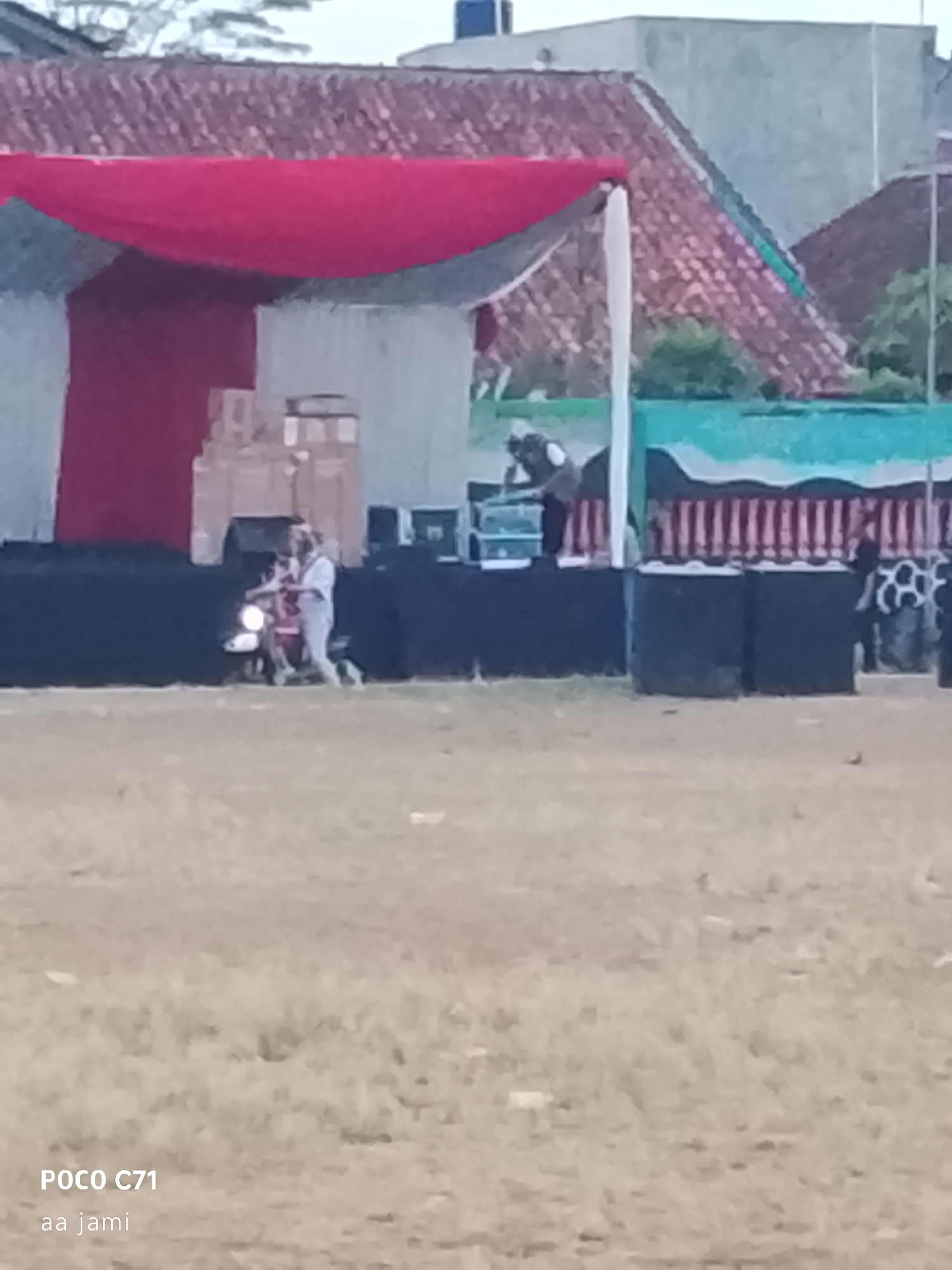 The image shows an outdoor event with a stage and a grassy field. The stage has a red canopy and is decorated with red and white fabric. There are speakers and some boxes on the stage. In front of the stage, two people are visible. One person is riding a motorcycle, and another person is standing next to it. The background shows buildings with red tiled roofs and some trees. The ground in the foreground is dry and covered with grass. A watermark at the bottom left corner of the image reads "POCO C71 aa jami". The overall scene suggests a cultural event or performance taking place in a public space, possibly in the city of Tasikmalaya, Indonesia, given the watermark.