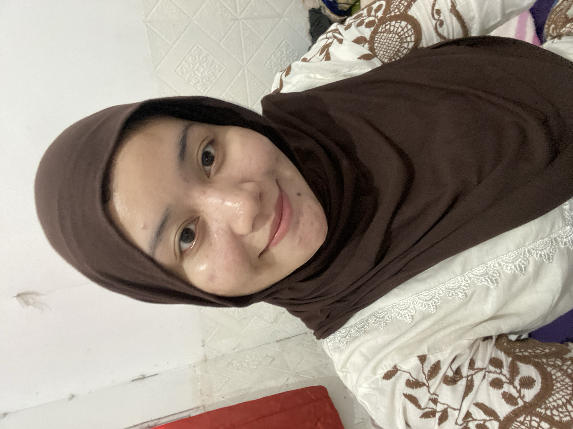 A young woman wearing a brown hijab and a white top with brown embroidery is taking a selfie. She is indoors, and the background appears to be a wall with a textured pattern. The lighting is somewhat harsh, creating highlights on her face. She has a mole on her chin.