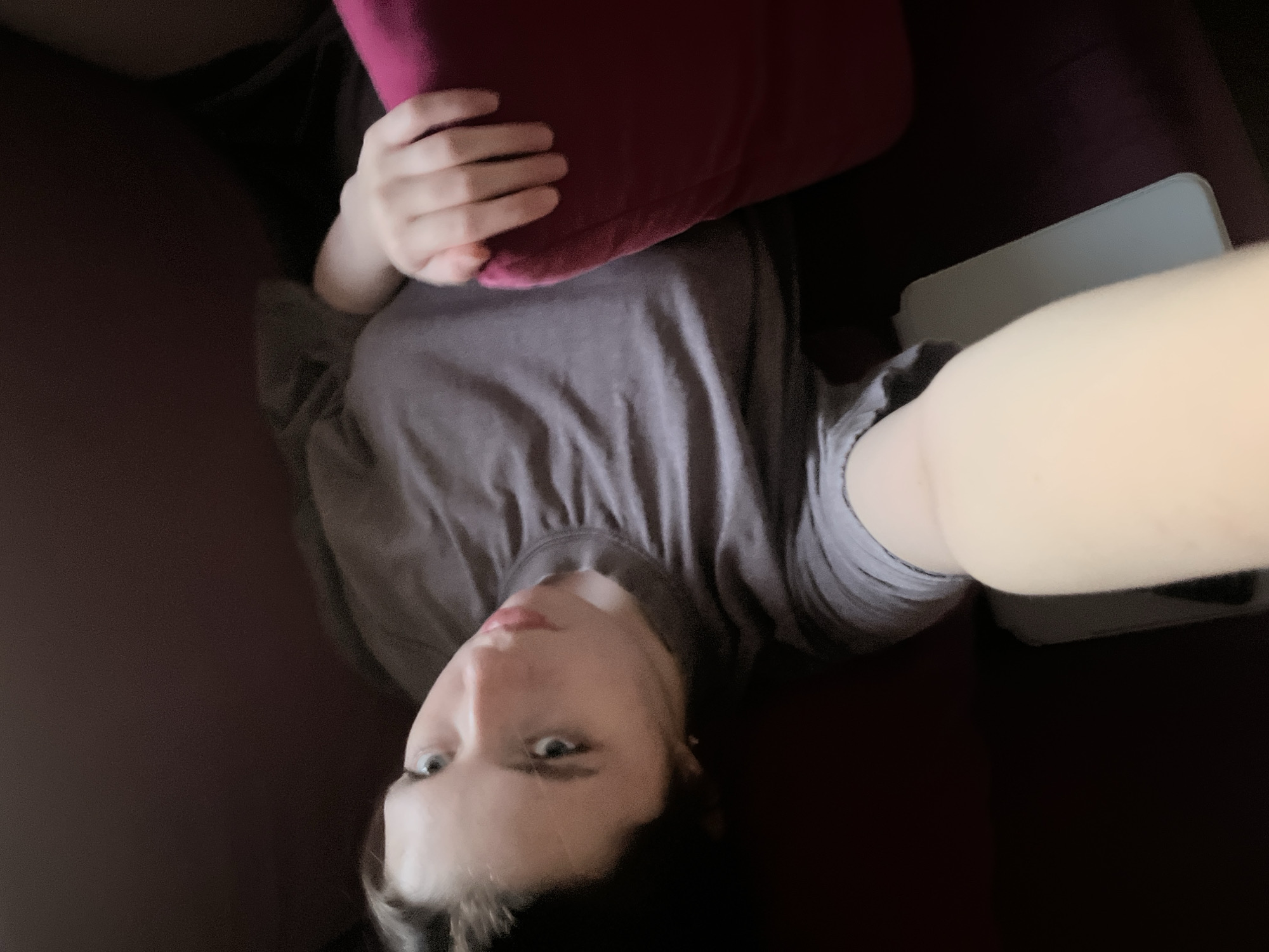 A person lies on a dark upholstered surface holding a red pillow, possibly on a couch or bed. The person wears a grey t-shirt. A white rectangular object is visible, possibly a tablet or phone. The location is unknown and there is no indication this photo was taken in Moscow, Russia.