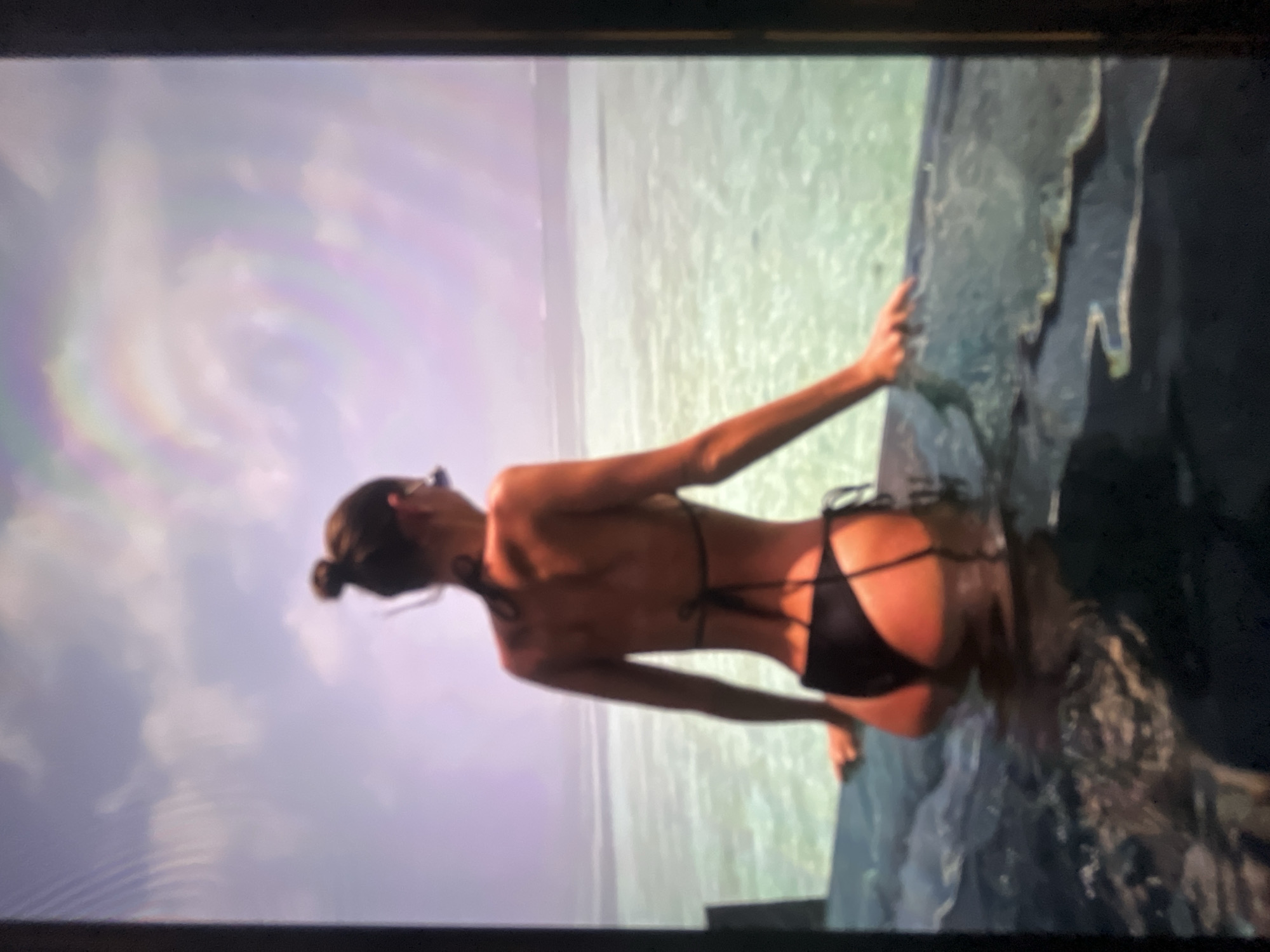 The image shows a person in a black bikini standing in clear turquoise water, facing away from the camera. The person has their hair pulled up in a bun and is wearing sunglasses. They are leaning on the edge of an infinity pool, with their hands resting on the edge. The water of the pool blends seamlessly with the ocean in the background, which appears to be a shallow, tropical lagoon. The sky is a light blue with some wispy clouds. The image is taken from a slightly elevated perspective. The location is not specified as Ravno pole, Bulgaria.