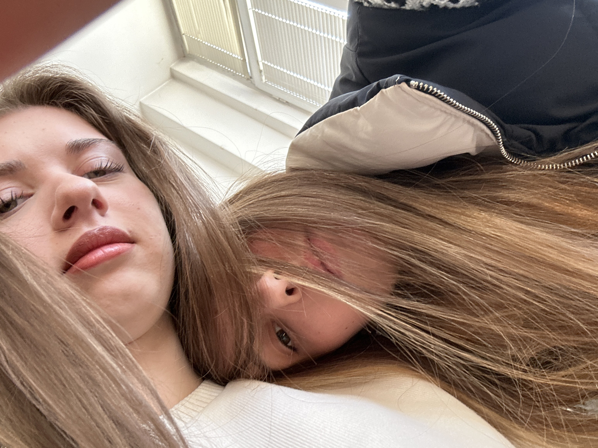 The image displays a close-up, low-angle shot of two young females, positioned intimately, likely for a selfie photograph. The individual at the top has long, straight, light brown hair partially obscuring her face, with visible eyes looking towards the camera and pink-toned lips. She wears a white, possibly ribbed, top. The second individual, positioned below and slightly behind, also has long, straight, light brown hair, which largely covers her face, with one eye and part of her mouth visible. She is wearing a dark (likely black or navy) puffer jacket with a silver-colored zipper and a light-colored (beige or light grey) inner collar lining. A patterned fabric, possibly checkered, is visible on the right side of her shoulder. Both subjects appear to be looking directly into the camera lens. The background is an out-of-focus indoor environment, revealing a white or light-colored wall/ceiling section and a window equipped with white horizontal blinds, partially open. No specific landmarks or architectural features are present to identify a precise location within Sofia, Bulgaria; the setting appears to be a generic interior space, possibly a classroom or residential room.