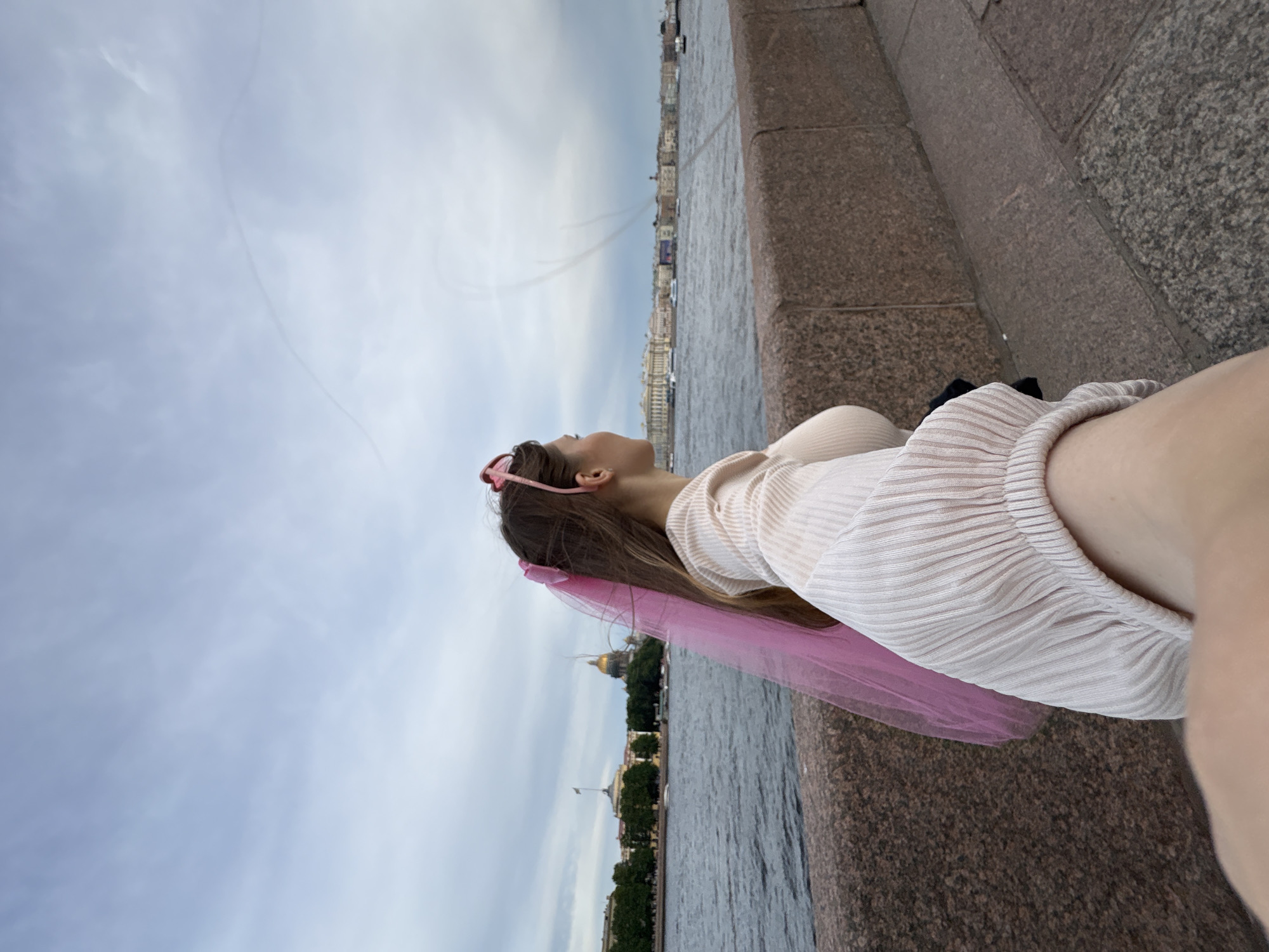 A woman, wearing a pink veil and a pale ribbed sweater, stands on a stone embankment overlooking a river in Saint Petersburg, Russia. Buildings, including a domed structure, line the opposite bank of the Neva River. The sky is cloudy, with faint contrails visible. The woman's face is turned to the right, looking out at the cityscape. The perspective suggests the image was taken from a first-person viewpoint, as if the viewer is holding the woman's hand.