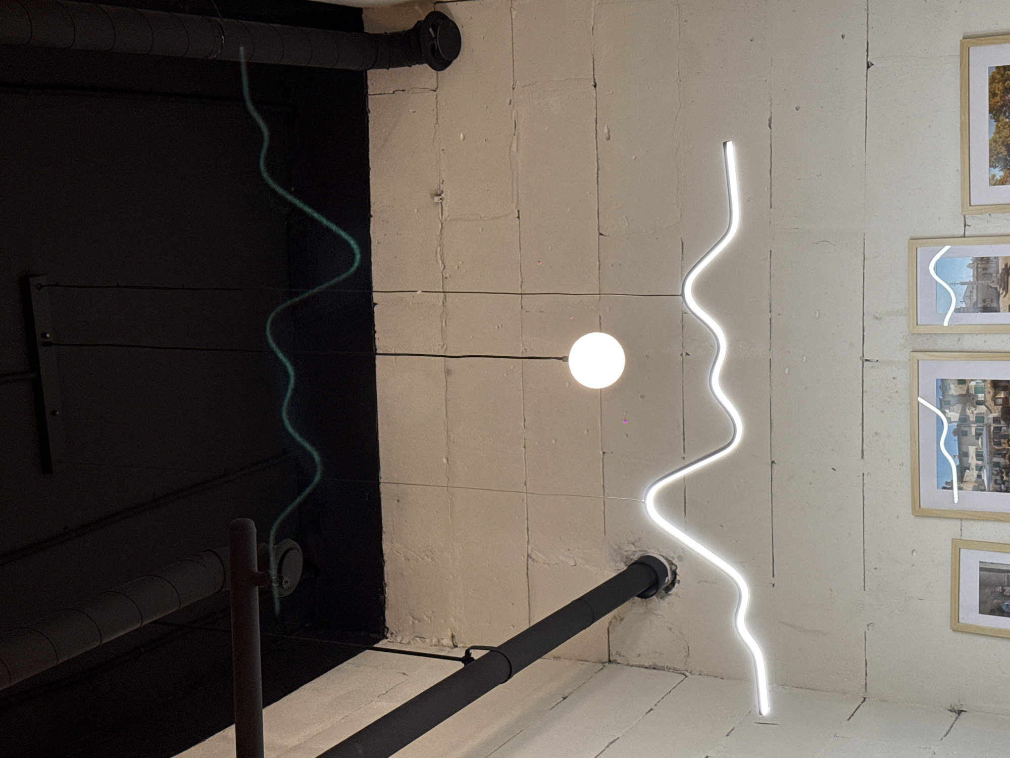 This vertical shot captures a modern indoor space, likely a commercial establishment, characterized by its industrial-chic aesthetic and unique lighting fixtures. The scene is dominated by two prominent, wavy LED light strips: one a vibrant green against a dark, possibly black, wall or ceiling section on the left, and another, bright white, against a textured, light-colored wall on the right.
A single spherical white light bulb hangs centrally, emitting a soft glow, with thin wires extending to it from above. Exposed black pipes, possibly part of a ventilation or plumbing system, are visible; one runs horizontally near the top of the frame, and another diagonally across the lower right, disappearing into the textured wall. The light-colored wall appears to be made of rectangular blocks or tiles, showing visible seams and imperfections.
On the right side of the frame, several framed pictures with light wooden frames are hung vertically on the textured wall. These pictures display urban or architectural landscapes, with one notably depicting buildings and another showing autumn foliage. The white wavy LED light fixture passes directly in front of these framed artworks, creating a visual overlay. A dark, possibly black, flat surface is visible on the left, contrasting sharply with the illuminated wall. In the lower left, a dark metal pole rises vertically. There are no people visible, and no specific activity is taking place. No text is discernible in the image. The artificial lighting suggests it could be evening or night, or an interior that relies solely on artificial light.