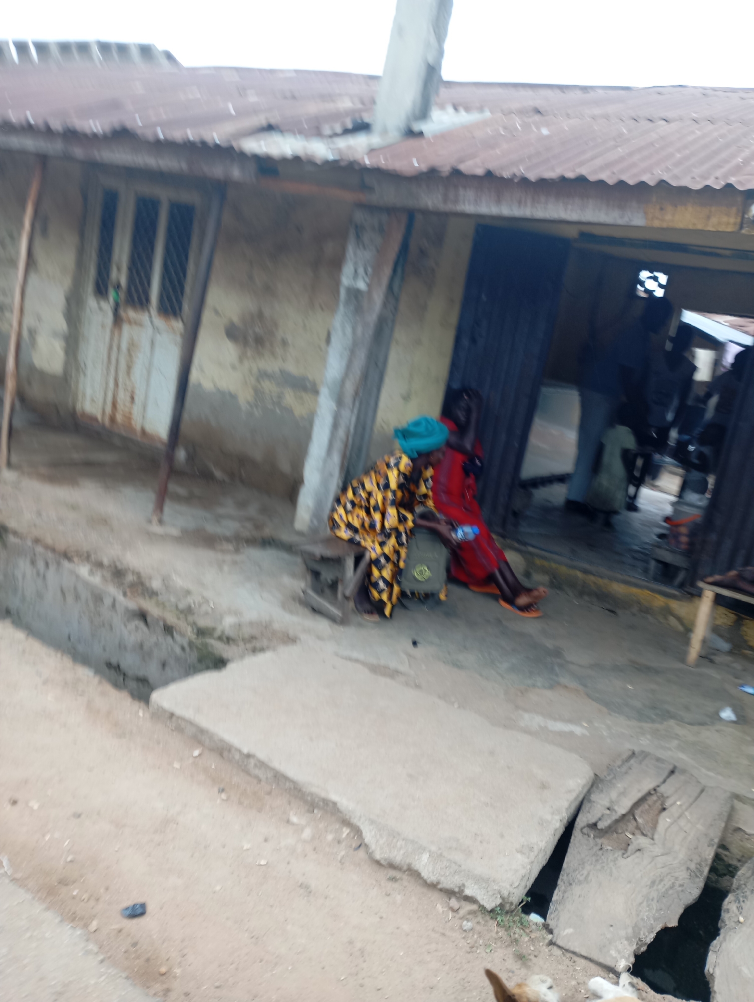 In Shendam, Nigeria, an outdoor scene captures a modest, weathered structure with a corrugated metal roof, likely a shop or dwelling. The walls appear to be concrete or stucco, showing signs of age and wear. In front of the building, two individuals are seated on a low platform or bench. One person, wearing a vibrant yellow and black patterned garment and a turquoise headscarf, is seated, leaning slightly forward. Next to them, another person in a red garment is also seated, their posture suggesting a moment of rest or conversation. A bottle of water and a backpack are placed on the ground near them. The ground in front of the building is a mix of dirt and concrete slabs, with a visible drainage channel running alongside. Further back, the entrance to the building reveals glimpses of interior activity, with other people partially visible within the darker space. The overall impression is a snapshot of everyday life in a built environment that reflects a rural or semi-urban setting, with utilitarian structures and the presence of people engaged in quiet activity.