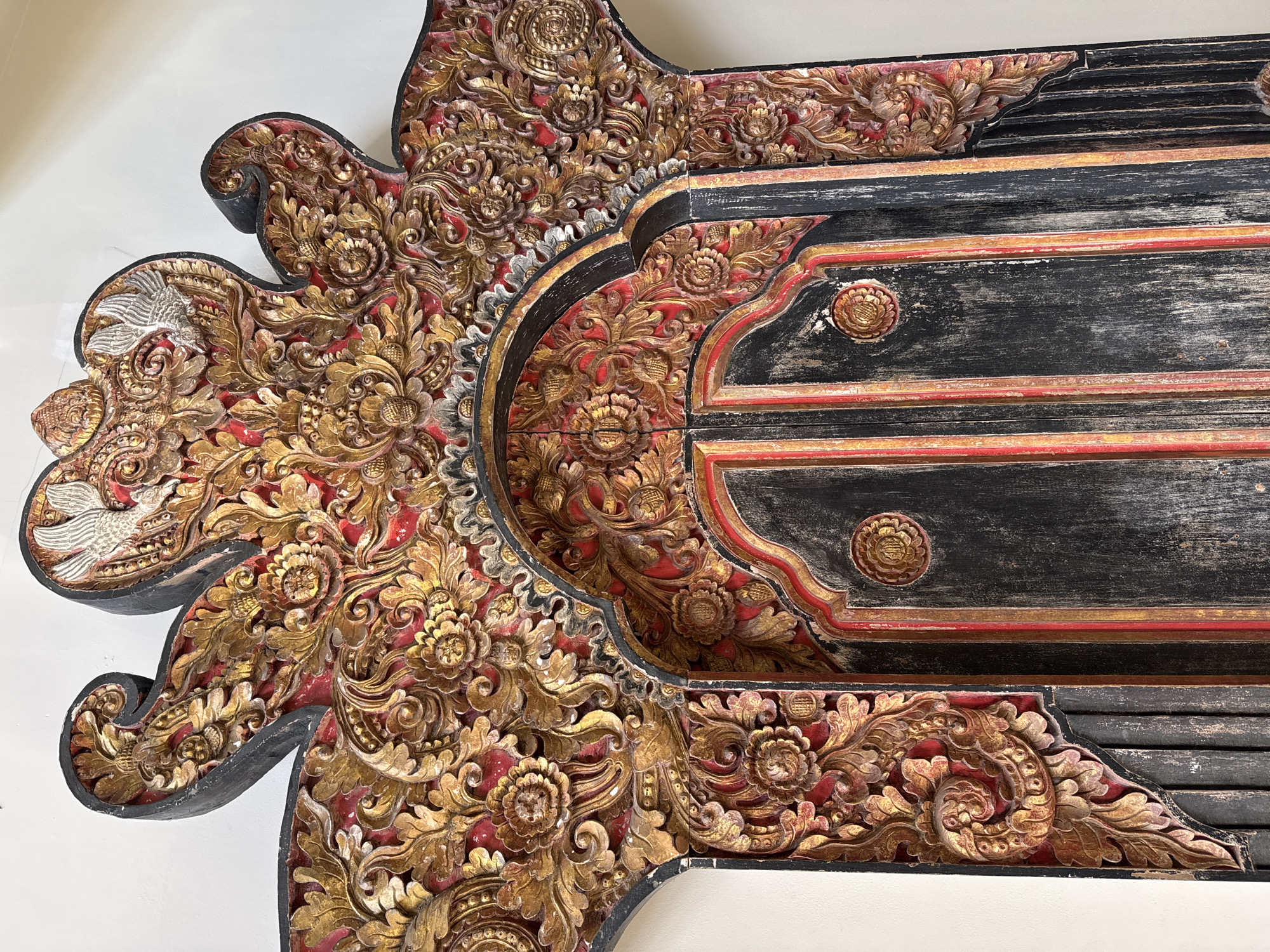 A detailed, ornate, carved wooden doorframe is depicted against a plain white wall in Denpasar, Indonesia. The doorframe is richly decorated with intricate floral patterns and scrollwork, primarily in gold and red hues. A black, distressed wood panel forms the actual door, with vertical red and gold trim. At the top of the doorframe, two silver bird-like figures are carved. The carving exhibits a high level of craftsmanship and appears to be an antique piece, likely adorning a traditional Balinese building or temple. The lighting is soft and even, highlighting the textures and details of the carving. There are no people or animals present in the image, only the static architectural element.
