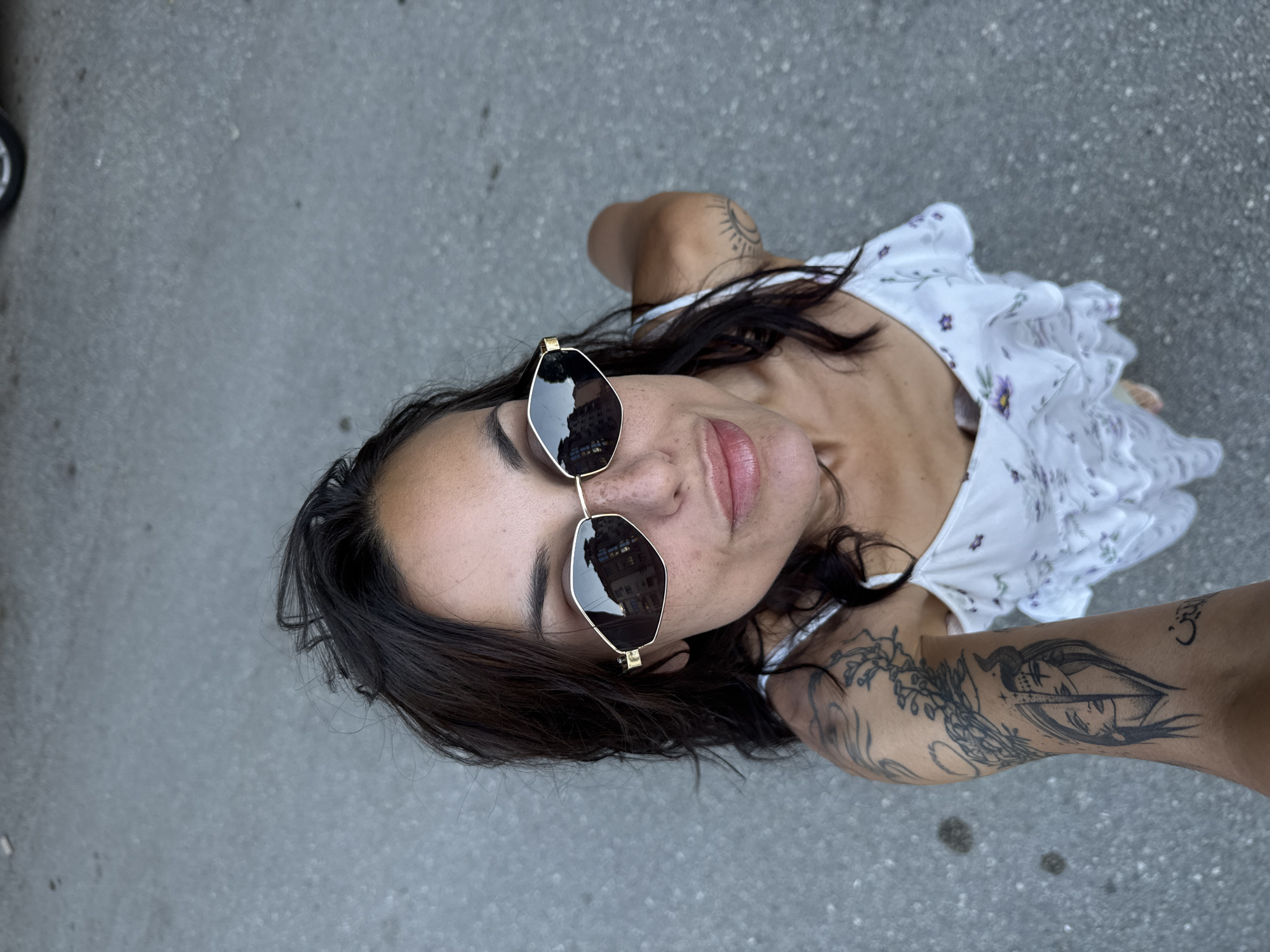 A woman with dark hair and sunglasses is captured in a high-angle shot from above, standing on an asphalt surface. She is wearing a white dress with a floral pattern and has multiple tattoos on her left arm, including a depiction of two faces and a sun and moon symbol. The sunglasses she wears reflect buildings. The background appears to be an urban environment, consistent with the city of Pleven, Bulgaria.