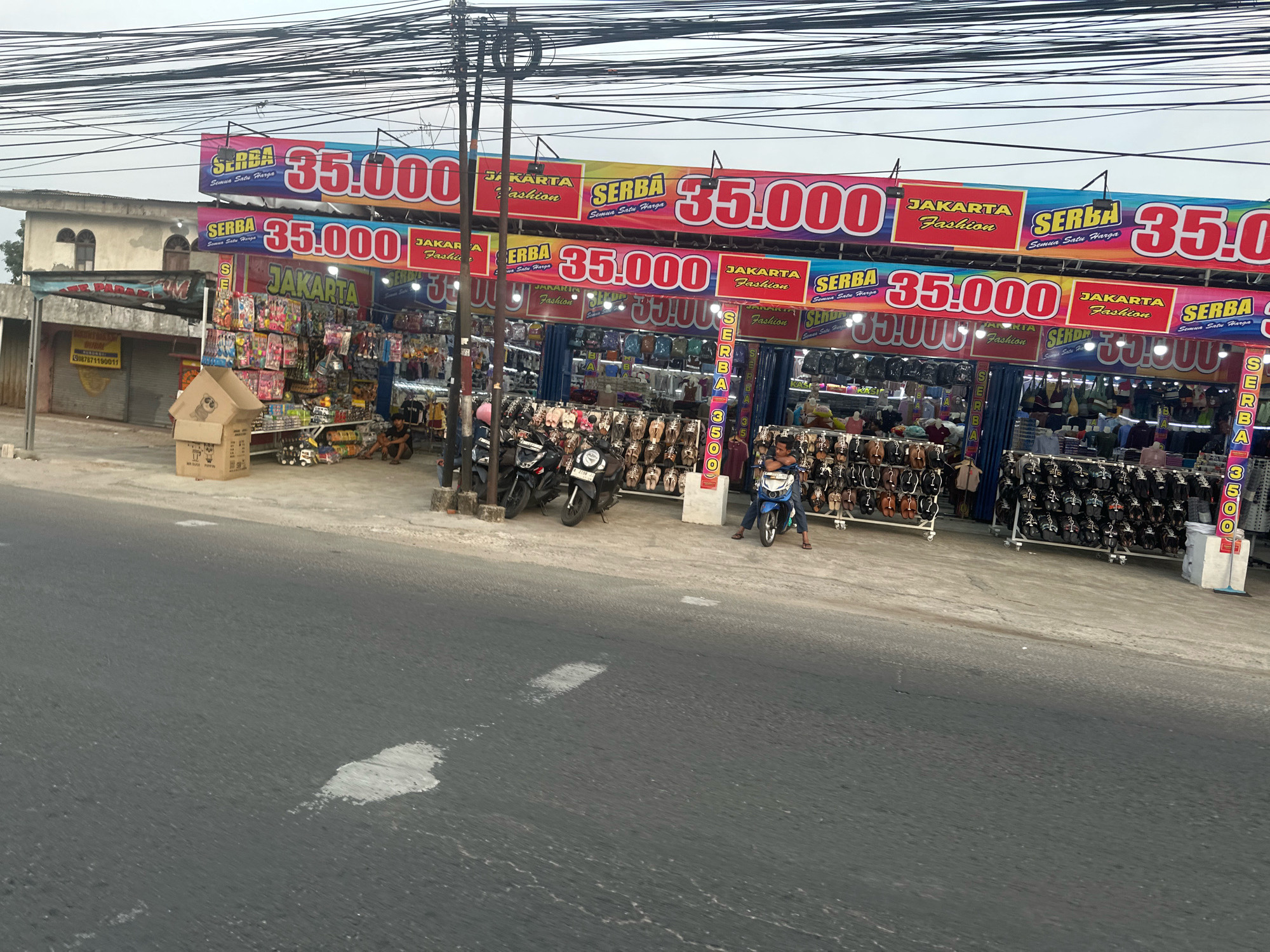 The image captures a street-level view of a row of shops with prominent banners advertising "SERBA 35.000" and "JAKARTA Fashion". The banners, in red and yellow, suggest a sale or a fixed price point for items sold in the shops. Several shops are visible, displaying merchandise like shoes, bags, and what appear to be toys or accessories. The shops are arranged along a sidewalk next to a paved road.
In the foreground, the road surface shows some wear and tear with patches of lighter color. A few motorcycles are parked in front of the shops, one blue motorcycle with a rider is a focal point. A person is seated on the blue motorcycle, wearing a dark shirt and light-colored pants, with their feet touching the ground. Another person is seen sitting on the ground near a pile of cardboard boxes.
The shops themselves have open fronts, allowing a clear view of the goods displayed on racks and shelves. The merchandise appears to be varied, including many pairs of footwear. The architecture of the buildings behind the shops is simple, with one building having arched windows. Electrical wires are strung across the top of the image, indicating an urban or semi-urban setting. The overall impression is that of a bustling commercial area, likely a marketplace or a street lined with retail outlets in Saruni, Indonesia.