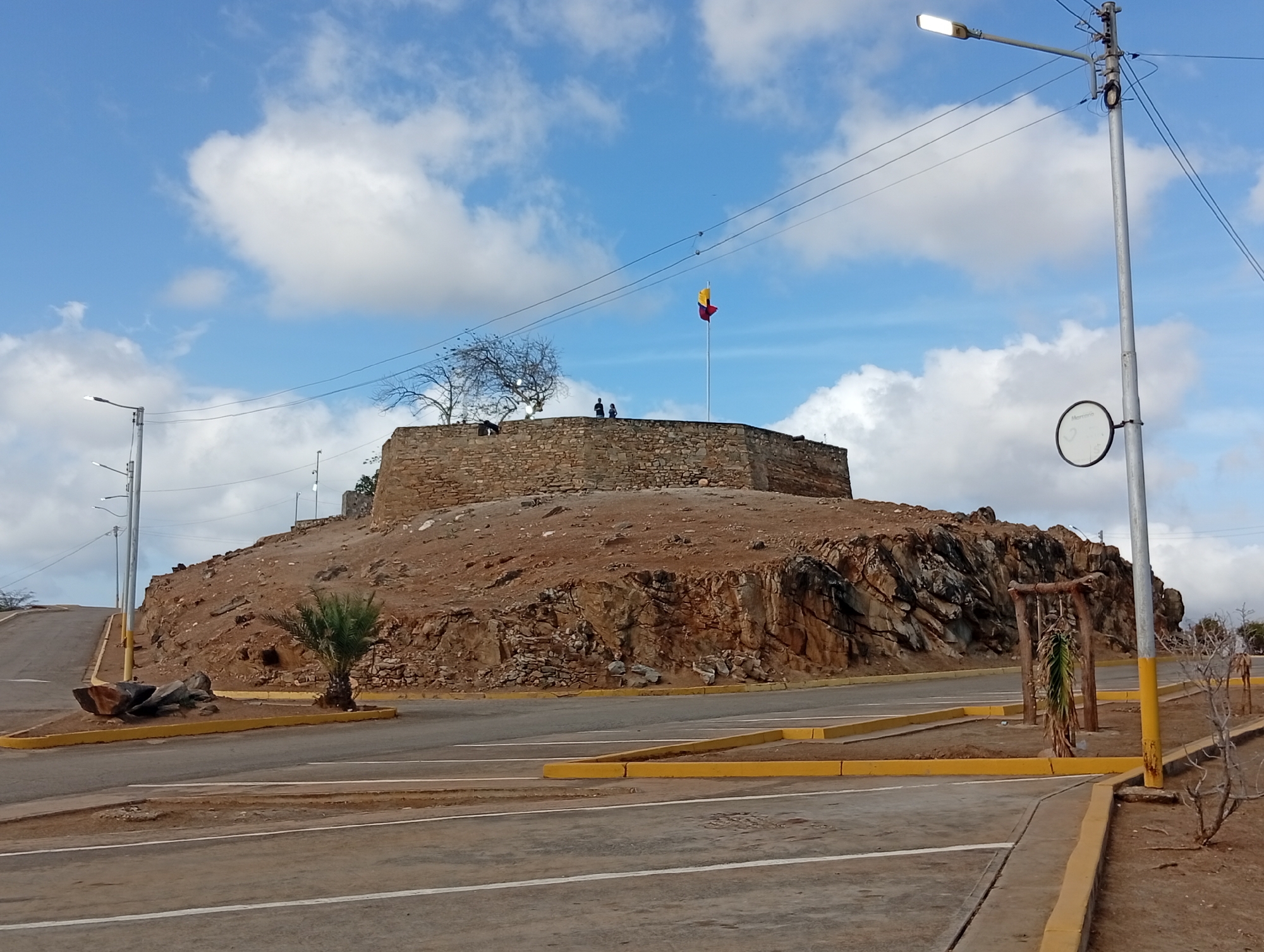 The image shows the Fortín de La Galera, a historical fortification located in Juan Griego, Venezuela. The fort sits atop a rocky hill with a stone wall and a flagpole displaying the Venezuelan flag. Two people are standing on the fort's wall. A bare tree is beside them.
The surrounding area includes paved roads, parking spaces delineated by yellow curbs, and lampposts. The foreground displays various roadside elements, including a small palm tree, a rock formation, and a makeshift wooden structure with decaying foliage.
The sky is blue with scattered white clouds. Powerlines are visible in the sky.