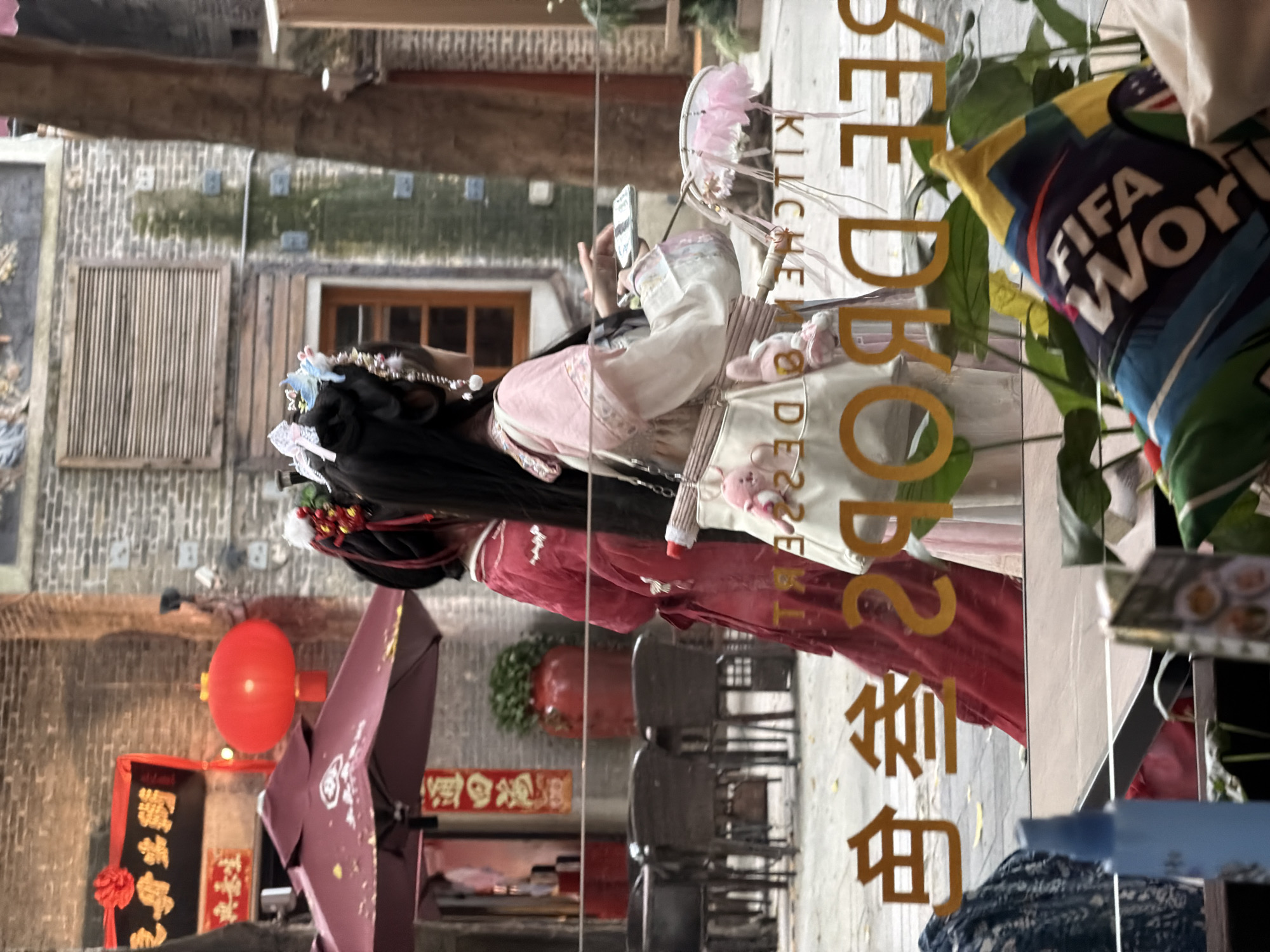 In Chancheng District, China, two individuals adorned in traditional Chinese attire are walking past a building with exposed brick walls. The person on the left wears a striking red garment, while the person on the right is dressed in a lighter pink and white outfit. Both individuals have elaborate hair decorations. In the foreground, a reflection on a glass surface reveals several yellow characters, possibly a sign, and a portion of a pillow with the "FIFA WORLD" logo. The background shows an outdoor seating area with tables and chairs, a large red umbrella, and a red lantern hanging from a building.