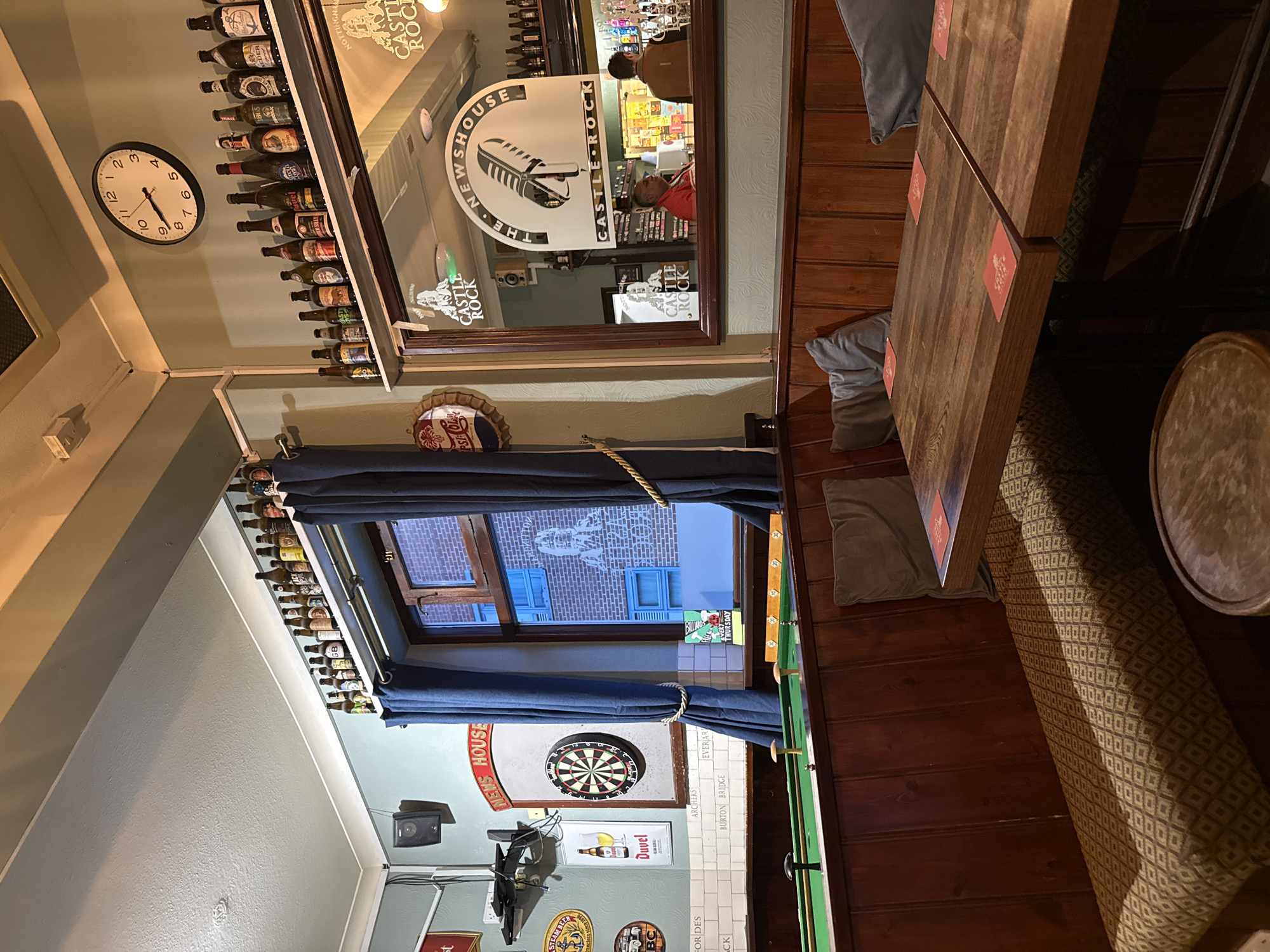 The image shows the interior of The News House pub in Nottingham, United Kingdom. The bar is located at the top of the photo, with bottles displayed on shelves. A mirror shows a partial reflection of the bar area, including two individuals. Below the bar, a clock on the wall reads approximately 6:51.
A window below the clock is partially covered by blue curtains, offering a view of the building exterior. Beneath the window, a dartboard hangs on the wall, along with several framed advertisements for alcoholic beverages and the Castle Rock brewery.
To the right of the window is a wooden table with two grey cushions, and a padded bench with a patterned fabric. A section of a pool table is visible on the right of the frame.
