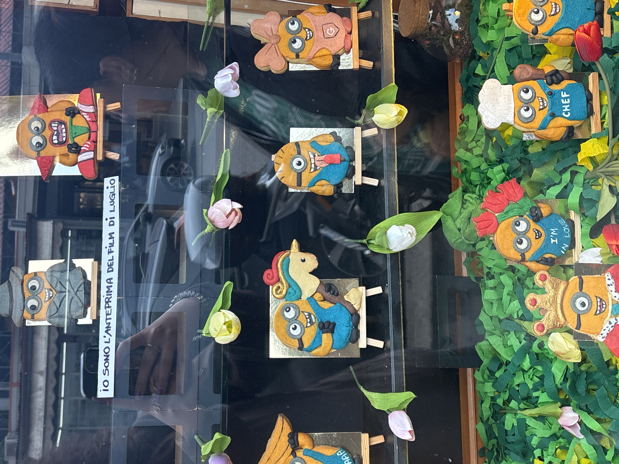 The image captures a display of handmade Minion-themed figures in what appears to be a shop window in Milan, Italy. The figures are crafted with felt and decorated to represent various Minion characters in different outfits and scenarios: a chef Minion, a king Minion, a Minion in love, and a Minion as a unicorn. These figures are positioned on miniature wooden stands. Artificial flowers, primarily tulips, are interspersed throughout the display. A sign in Italian, "Io sono l'anteprima del film di Luglio" (I am the preview of the July film), can be seen, suggesting a promotion for a movie. The background reflects the urban environment of Milan, and a partial reflection of a person and a parked car can be observed in the window. The display is arranged against a black background, emphasizing the colorful figures and flowers. The display is positioned in a window of what looks like a shop.