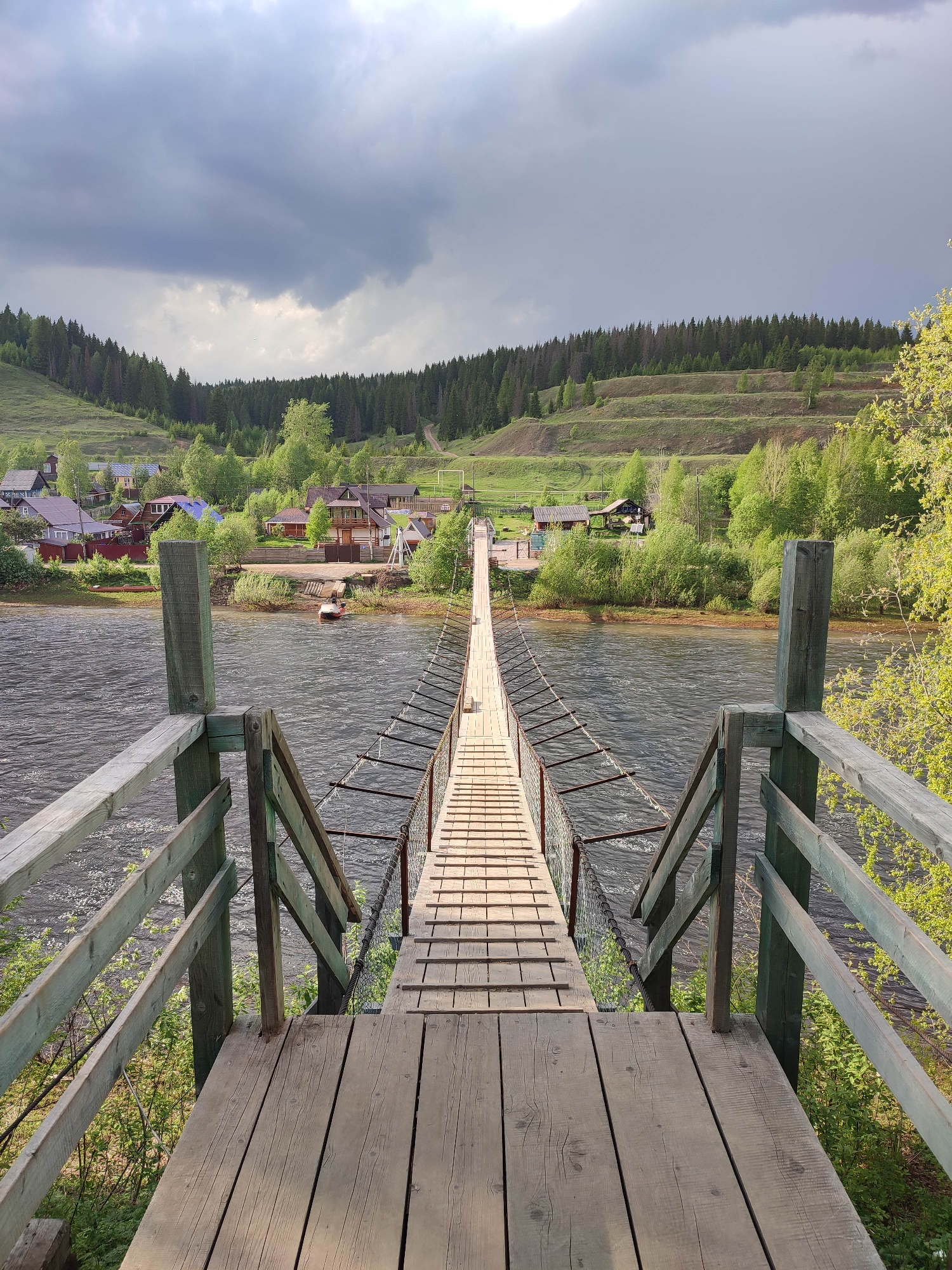 This image depicts a suspension bridge over a river, leading to the village of Усьва in Russia. The bridge is constructed of wooden planks and supported by metal cables with wooden railings on either side leading to it. The river is wide and flowing. In the background, there's a village with houses interspersed among green trees. A terraced hillside rises beyond the village. The sky is overcast with storm clouds.
