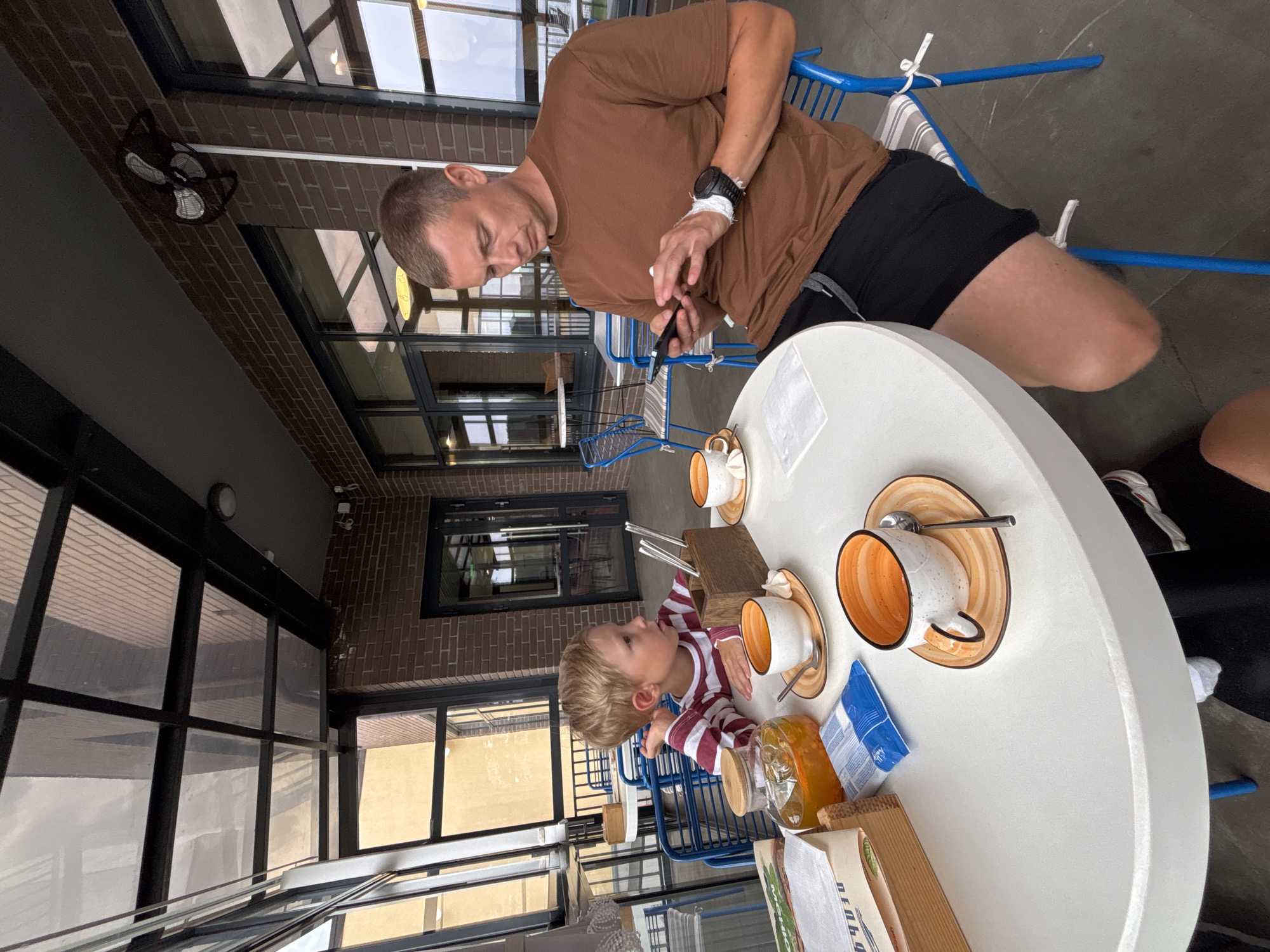 The image depicts an outdoor seating area at a cafe, possibly in Chaikovsky, Russia. A man in a brown t-shirt and black shorts is seated at a white round table, looking at his phone. He has a watch on his left wrist. In front of him is a cup of coffee and a small white pastry. A young boy with blond hair, wearing a striped long-sleeved shirt, is seated opposite him. He is looking towards the man. On the table between them are two cups of coffee, a carafe of orange-colored drink, a small package, and a wooden holder with cutlery. The background shows brick walls, large windows, and a transparent roof. Blue chairs are visible around the table.