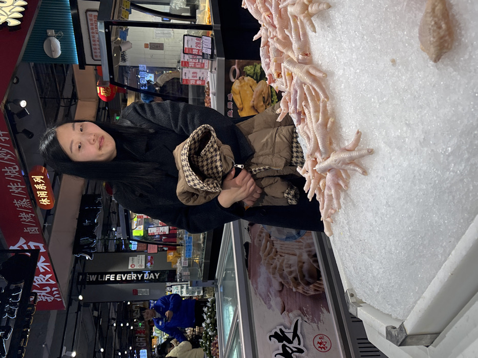 The image displays an indoor scene within a supermarket located in Jiangjin, China. In the immediate foreground, a white refrigerated display bin holds a large pile of raw, light-pink chicken feet resting on a bed of crushed ice. A single raw chicken wing is also visible on the ice in the lower right. Centered in the midground, a woman with long, dark hair, wearing a black long-sleeved coat and a black turtleneck, stands holding a folded brown quilted jacket with a brown and white houndstooth-patterned lining. She faces slightly to her right with a neutral expression. Overhead, red banners with white Chinese characters are visible, including "熟食" (Cooked Food) and "休闲系列" (Leisure Series), along with various cooking methods. To the left in the background, a man in a blue uniform shirt is visible, looking at his mobile phone while standing near another refrigerated section. To the right, behind the woman, a counter displays various packaged food items with price tags, and a red Chinese lantern hangs above. A black column with "NEW LIFE EVERY DAY" in white text is positioned in the midground on the left. The environment is well-lit with overhead lighting, consistent with a modern retail food establishment.