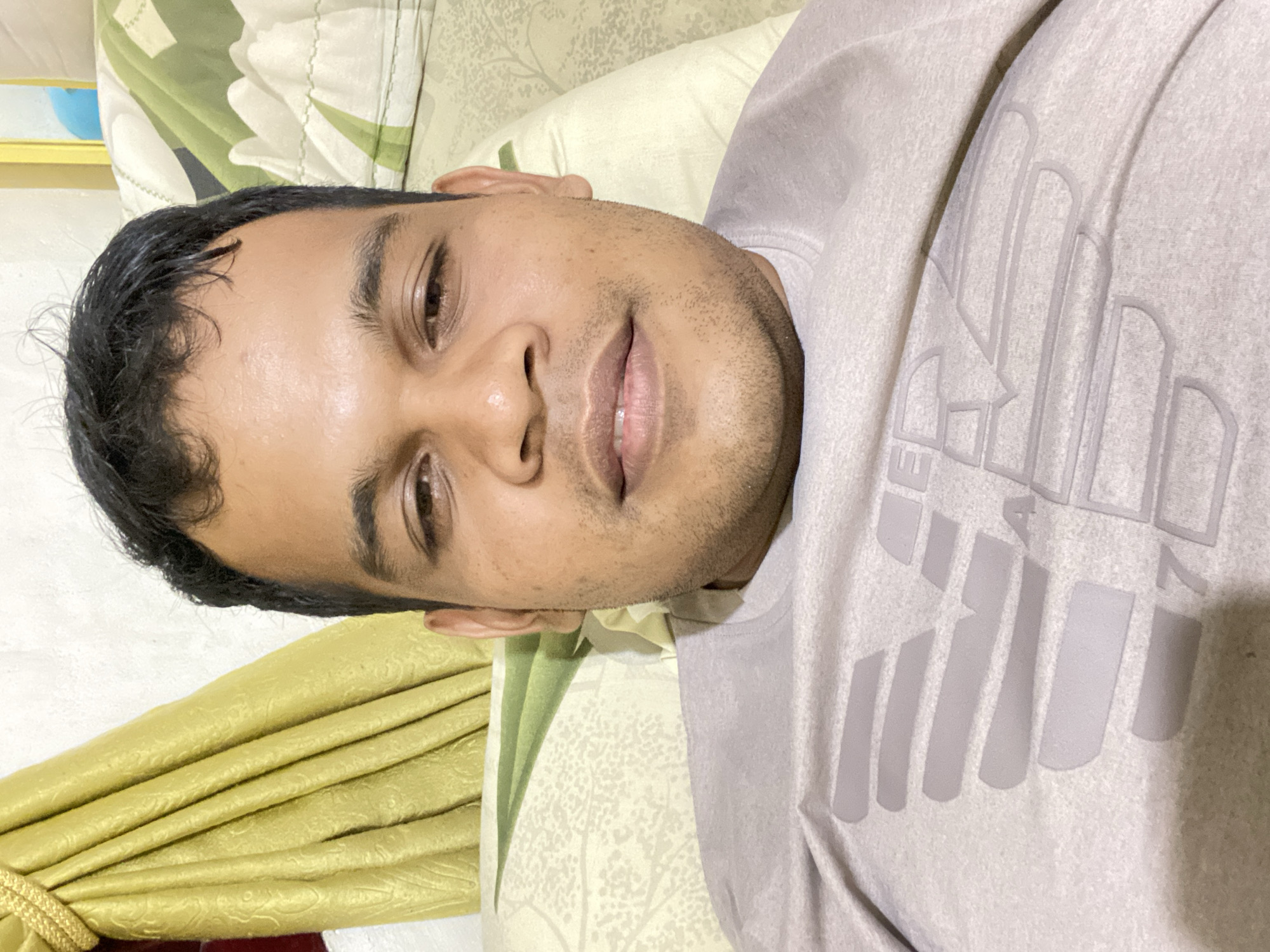 A close-up, selfie-style photo shows a man with dark hair and light brown skin resting on his back. He is wearing a light purple t-shirt with a grey eagle logo and the number "7" visible on the front. The man is lying on a bed with a light-colored patterned comforter and a green and white patterned pillow. A gold curtain is visible in the background on the left side of the image.