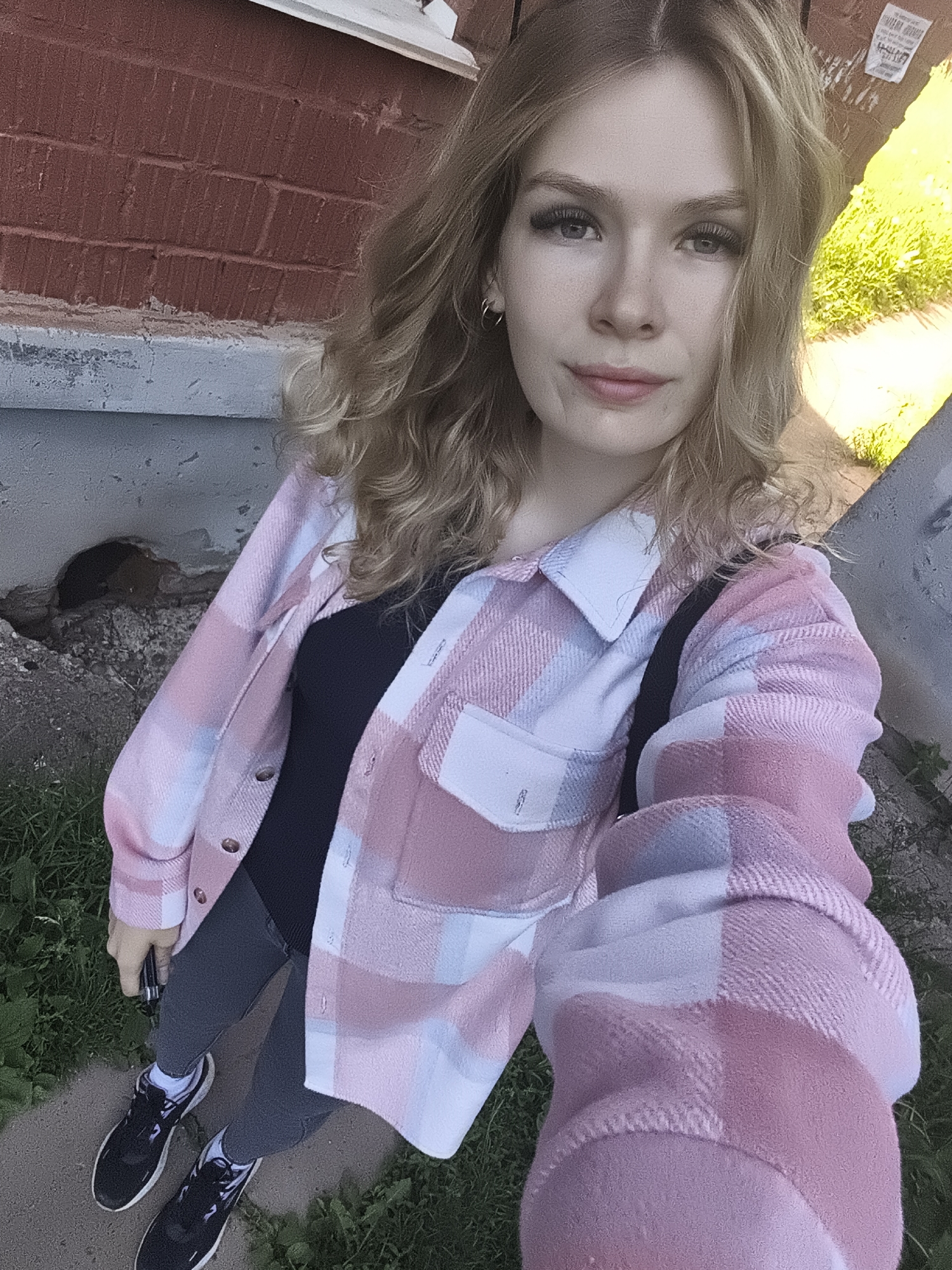 A young woman is captured in a full-body selfie, looking directly at the camera with a neutral to slightly smiling expression. She has long, wavy blonde hair, light-colored eyes with prominent eyelashes, and light makeup. She is wearing a casual outfit consisting of an oversized, long-sleeved plaid shirt or jacket in shades of pink, white, and light blue, over a black top. Her lower body is dressed in dark grey or black form-fitting pants, white socks, and black sneakers with white soles. A black strap, likely from a bag, is visible over her left shoulder. She holds a small, dark device, possibly a phone, in her right hand, which is positioned near her hip.
The setting appears to be outdoors, likely in an urban or suburban environment during daytime, indicated by the bright natural light. The background features a red brick wall, a concrete ledge or path, and patches of green grass. There's a dark, irregular hole visible in the concrete near the base of the wall. On the brick wall in the upper right background, there are remnants of a white paper or poster with indistinct, illegible text. No specific weather conditions are overtly visible, but the lighting suggests clear skies.
The activity is a self-portrait or selfie, evident from the camera angle and the subject's direct gaze. The overall impression is casual and everyday.
(Location context provided: Izhevsk, Russia)