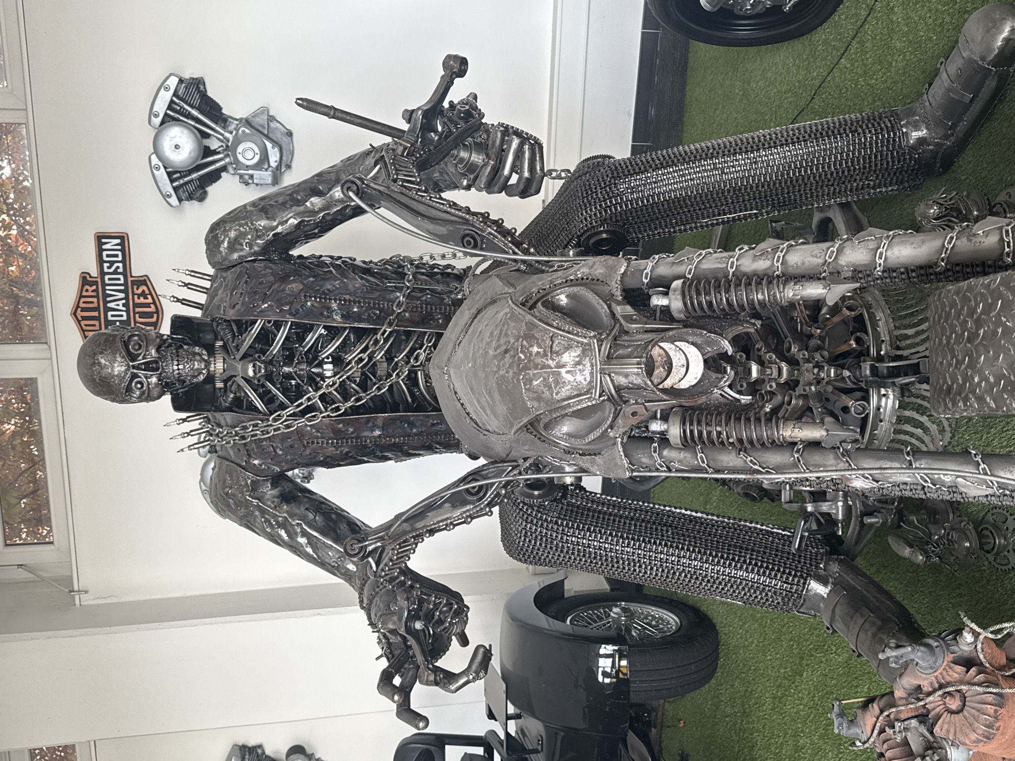 The image shows a metal sculpture of a skeleton riding a motorcycle. The sculpture is made from various metal parts, including chains, gears, and engine components. The skeleton is posed as if riding the motorcycle, with its hands gripping the handlebars. The sculpture is located indoors, possibly in a garage or workshop. A Harley-Davidson sign and engine part adorn the wall. There is a black vehicle and artificial green grass in the background. The location is specified to be in Bietigheim-Bissingen, Germany, but there are no visual cues in the image to confirm the location.