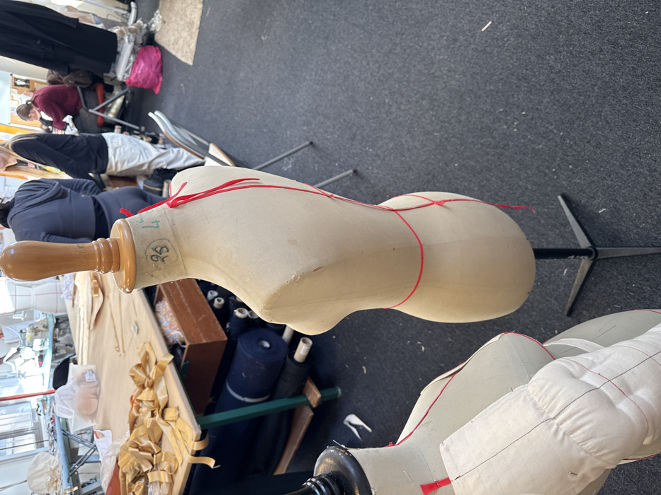The image displays an interior view of a fashion or textile workshop, identified as being located in Arcueil, France. The primary subject in the immediate foreground is a beige fabric dress form mounted on a black metal tripod base, oriented slightly towards the left. This dress form features a wooden finial at the neck, which is marked with "95" and "L" (or "4"). Red string or tape is meticulously draped around the dress form, outlining contours from the neck, over the shoulders, down the back, and around the waist. To the lower left, a partial view of a second beige fabric dress form is visible, distinguished by red stitching delineating bust lines and what appears to be an attached white fabric element, possibly a sleeve or padded panel.
The floor throughout the visible area is covered with dark grey carpeting. In the midground to the left, a light-colored wooden work table with green metal legs is situated, laden with numerous cut fabric pieces, predominantly tan or gold in hue, alongside various workshop materials. Beneath this table, rolls of dark-colored fabric are stacked on shelves. The background reveals several individuals in various states of activity within the workshop: one person, seen from the back, wears a dark grey hoodie and dark pants; another individual with blonde hair, wearing a black top and light-colored pants, stands partially visible to the right of the first person; and a third person, dressed in a dark red top, is seated at a table further back on the right. A silver folding chair and a prominent bright pink plastic bag are also present in the background on the right side of the frame. The workshop space is brightly illuminated, suggesting ample ambient light.