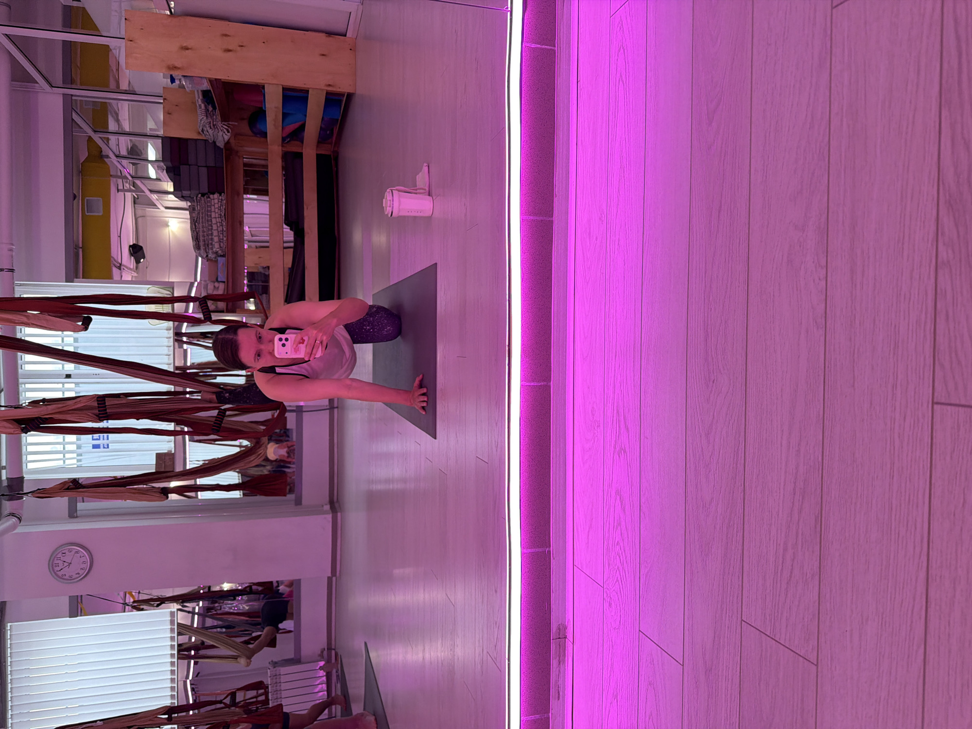 The image shows a woman in a yoga studio performing a yoga pose on a mat. She is holding a smartphone in her left hand, taking a selfie. The studio has wooden floors and aerial yoga hammocks hanging from the ceiling. A pink LED light strip runs vertically along the right side of the image, illuminating the wall. In the background, there are shelves with yoga props and equipment, as well as a clock on the wall. The overall lighting is dim, with the pink light casting a vibrant hue. There are no visible signs of time of day or weather, but the setting suggests an indoor recreational activity. The woman's expression is neutral, focused on her pose and the selfie. No text is visible in the image.