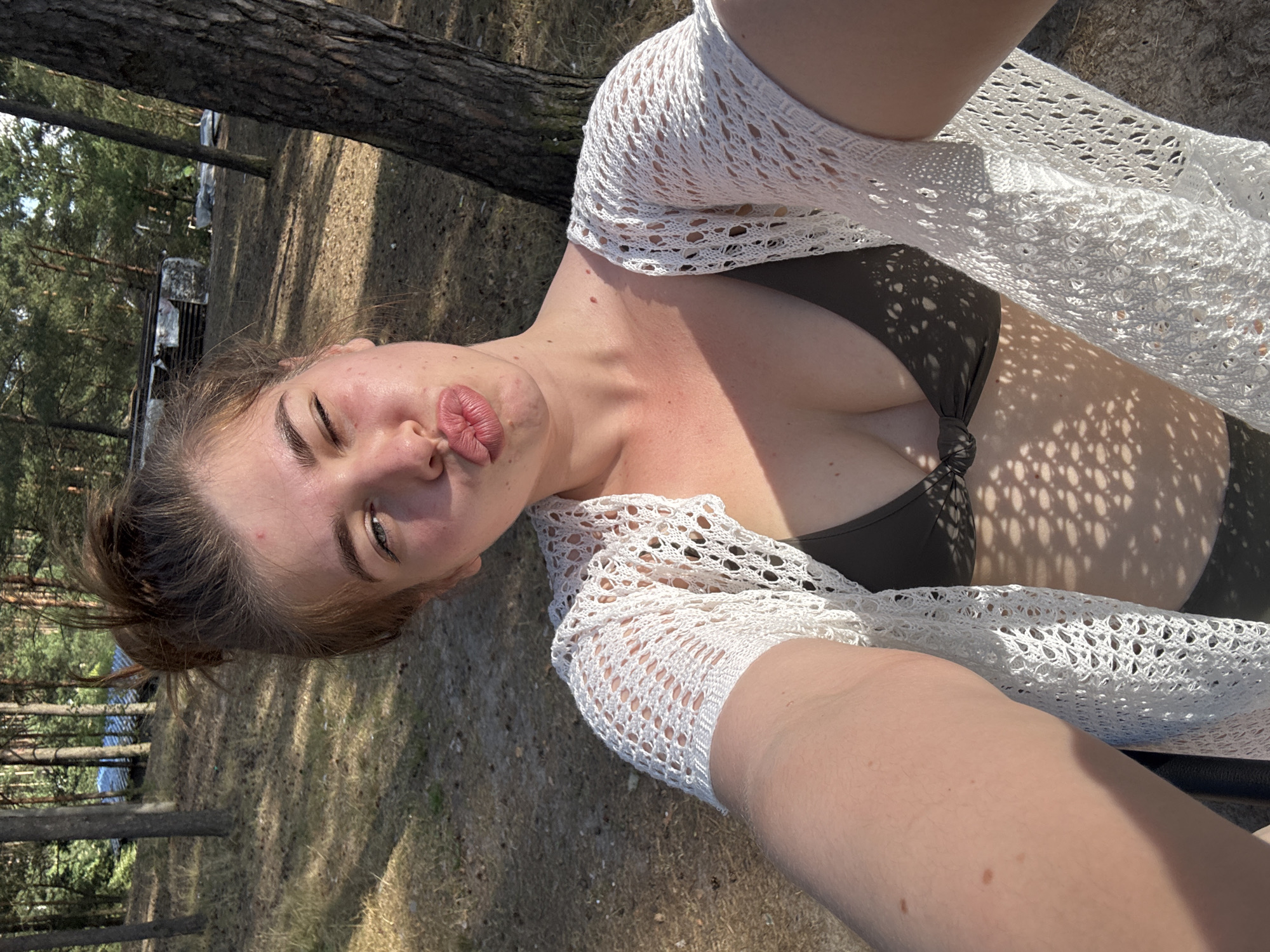 A young woman with her hair in a messy bun is taking a selfie outdoors in what appears to be a wooded area with sandy ground and dry grass. She is wearing a dark-colored bikini top and a white, open-knit cardigan. Her face is turned towards the camera with her lips pursed as if blowing a kiss, and one eye is partially closed. Sunlight casts dappled shadows on her and the surrounding environment. In the background, trees are visible, and some debris or structures can be seen through the trees. The overall impression is a casual, sunlit moment in a natural setting.