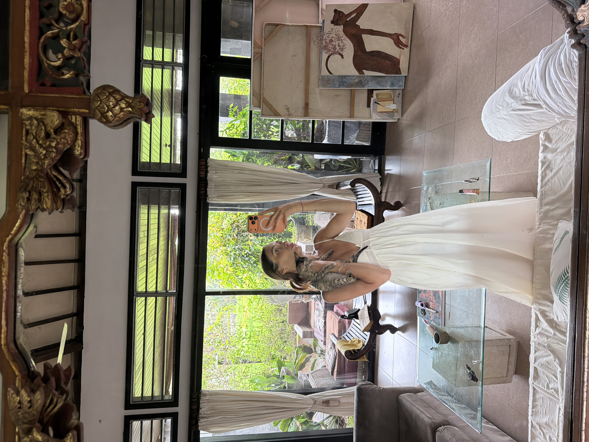 A woman is taking a selfie in the reflection of a large mirror or window, holding a small tabby kitten in her arms. She is wearing a white dress and holds a smartphone with a prominent camera bump.
The interior visible in the reflection features a glass-topped coffee table with several small items, a grey sofa, and a bed with white bedding, including a pillow adorned with a green leaf design. Against a wall, multiple artworks are stacked, one prominently featuring a stylized reddish-brown human figure. An ornate dark wooden chair with a yellow cloth is also visible. The floor is tiled.
Beyond the large windows, a vibrant, lush green tropical landscape is visible, suggesting an outdoor garden or patio area with dense foliage and what appears to be outdoor seating. The windows have dark frames and are partially draped with white curtains.
The bright natural light indicates it is daytime. The blend of ornate wooden decor and tropical greenery strongly suggests the specified location of Tegallalang, Indonesia. The woman's expression is focused on her phone, and the kitten appears calm. No discernible text is visible on any objects.
