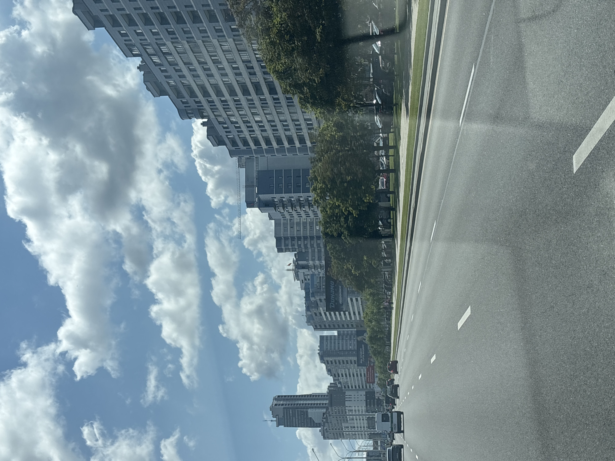 The image shows a wide, multi-lane asphalt road in a city. Several cars are visible on the road, traveling in the same direction. On the right side of the road, there are tall, modern apartment buildings with many windows, interspersed with green trees. A pedestrian walkway and parking area with more cars are visible between the road and the buildings. The sky is blue with fluffy white clouds. A tall skyscraper is visible in the distance on the left side of the image. Signs are visible on the buildings, one of which reads "DYNAMO". This scene is characteristic of a modern urban environment in Minsk, Belarus, specifically along one of the major avenues.