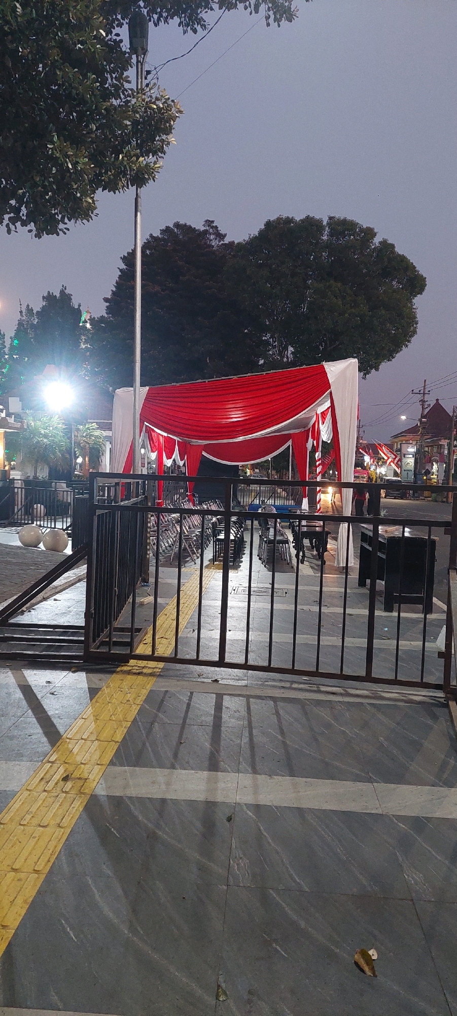 This image shows an outdoor event setup in Lumajang, Indonesia, likely in front of a building or public space. A large red and white tent, possibly for a celebration or gathering, is erected. Rows of empty chairs are neatly arranged beneath the tent, suggesting an upcoming event. In the foreground, a black metal fence separates the viewer from the seated area. The ground is paved with gray tiles and a yellow tactile paving strip, indicating a pedestrian walkway. A street with a few vehicles and buildings can be seen in the background. The overall atmosphere appears calm, with the event setup ready for use.