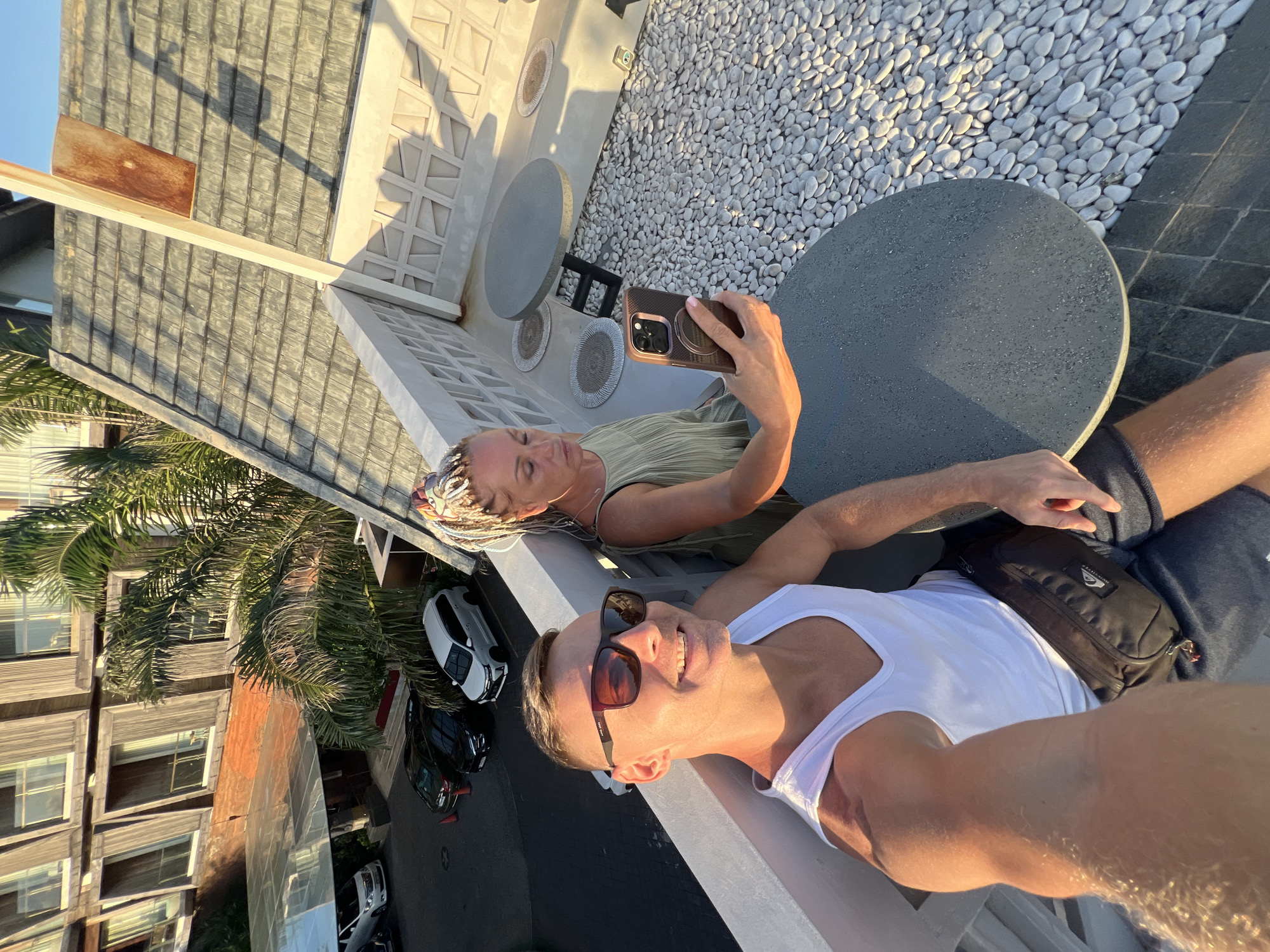 Two individuals, a man and a woman, are positioned on a rooftop in what appears to be Kuta, Indonesia, based on the architectural style and presence of palm trees. The woman is holding a smartphone, likely taking a photo or video of the pair. They are near a small, round table placed on a rooftop terrace adorned with white pebbles and decorative wall elements. In the background, a multi-story building is visible, with several cars parked on the street. The lighting suggests it is daytime, potentially late afternoon or early evening. The man is wearing sunglasses and a white tank top, while the woman is in a neutral-colored, sleeveless top. He is tanned and has a black shoulder bag. The woman has blond braids.