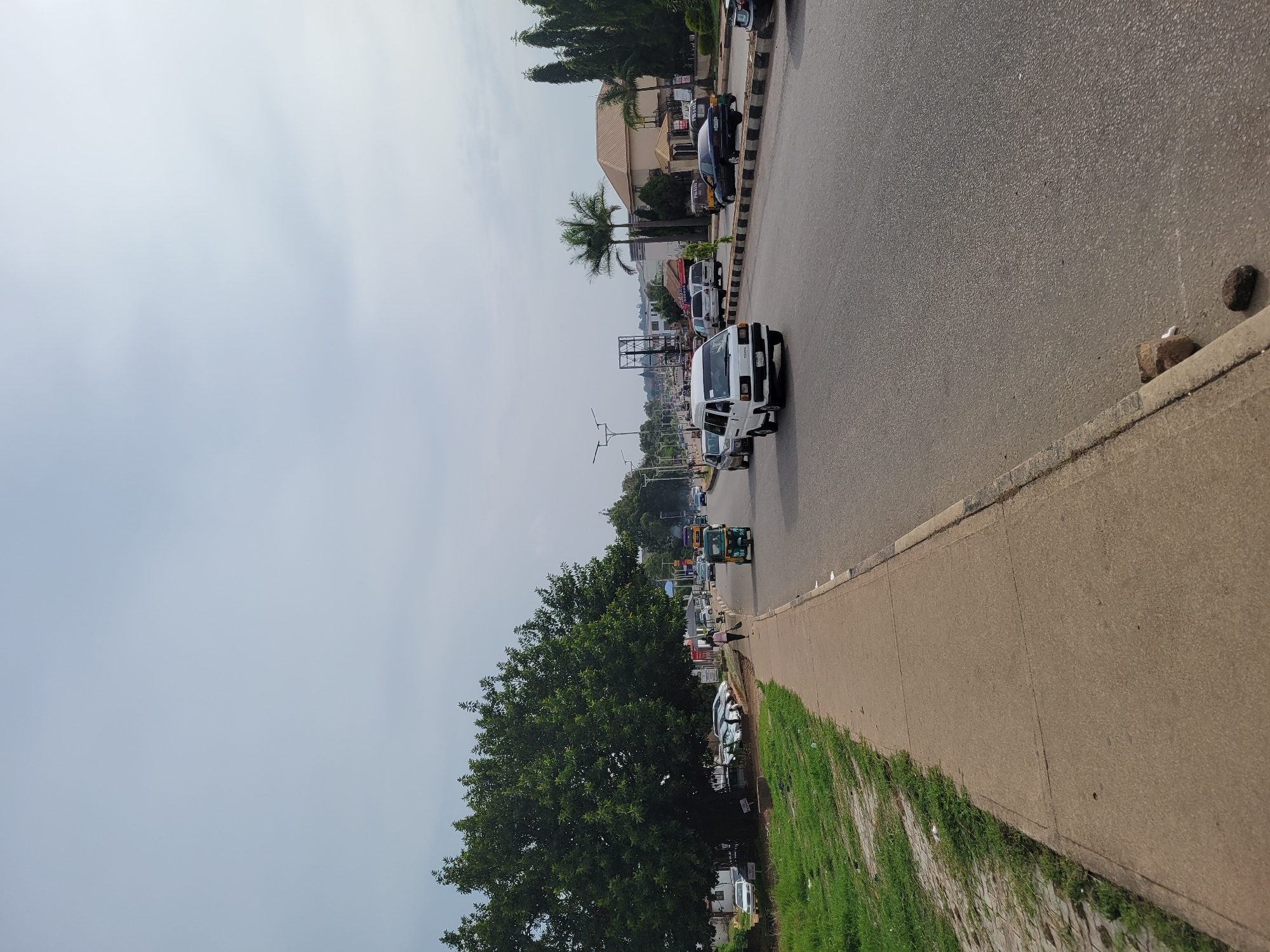 The image presents a daytime street scene, likely in an urban or semi-urban environment. The perspective is from the side of a road, looking down a street lined with buildings and vegetation.
On the right side of the image, there's a broad asphalt road, upon which several vehicles are moving or parked. In the foreground, a white van is prominent, facing away from the viewer. Further down the road, various cars, trucks, and a colorful motorized rickshaw (often called a "keke napep" in some regions) can be seen. On the left side of the road, more cars are parked, and the road is bordered by a black and white striped curb.
Adjacent to the road on the right is a wide concrete sidewalk, which has patches of green grass growing alongside it and further into the foreground. Scattered rocks are visible on the ground near the edge of the sidewalk. A large, leafy green tree dominates the midground on the right, and other trees, including tall palm trees, are visible further down the street on the left. A few pedestrians are visible on the sidewalk, engaged in walking.
In the background, behind the trees and parked vehicles, several buildings with varied architectural styles can be observed, suggesting a mix of commercial and residential structures. Street light poles and overhead lines crisscross the view. The sky occupies a significant portion of the image, appearing bright but overcast or hazy, with a uniform light blue-grey color, indicating a diffused light source.
The activity depicted is typical daily street life, with traffic flow and pedestrian movement. The time of day is clearly daytime, characterized by bright, diffuse lighting and no strong shadows. The presence of palm trees and the specific type of motorized rickshaw hint at a tropical or subtropical setting, possibly in West Africa. No discernible text is visible on any signs, vehicles, or buildings.