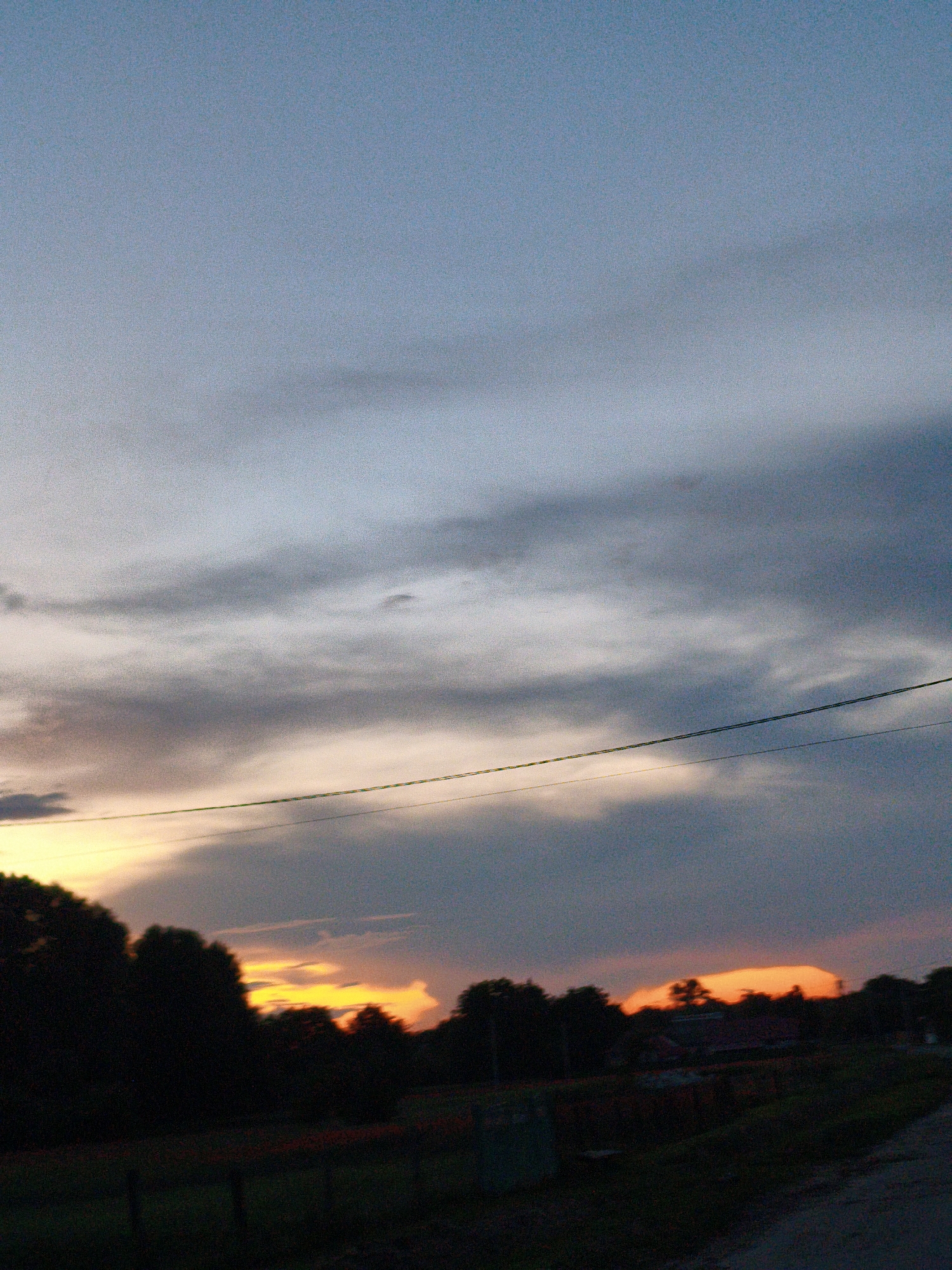 The image depicts a rural landscape at dusk. The sky is a gradient of blue and grey, with streaks of pale orange and yellow visible near the horizon, indicating the setting sun. Silhouetted against the colorful sky are dark trees and the roofs of a few buildings. A power line stretches horizontally across the middle of the frame. The foreground is dark and out of focus, suggesting it is evening or the camera is focused on the distant scene. The image captures a serene, twilight atmosphere. There is no indication of the city of Ustyvytsia, Ukraine within the image.