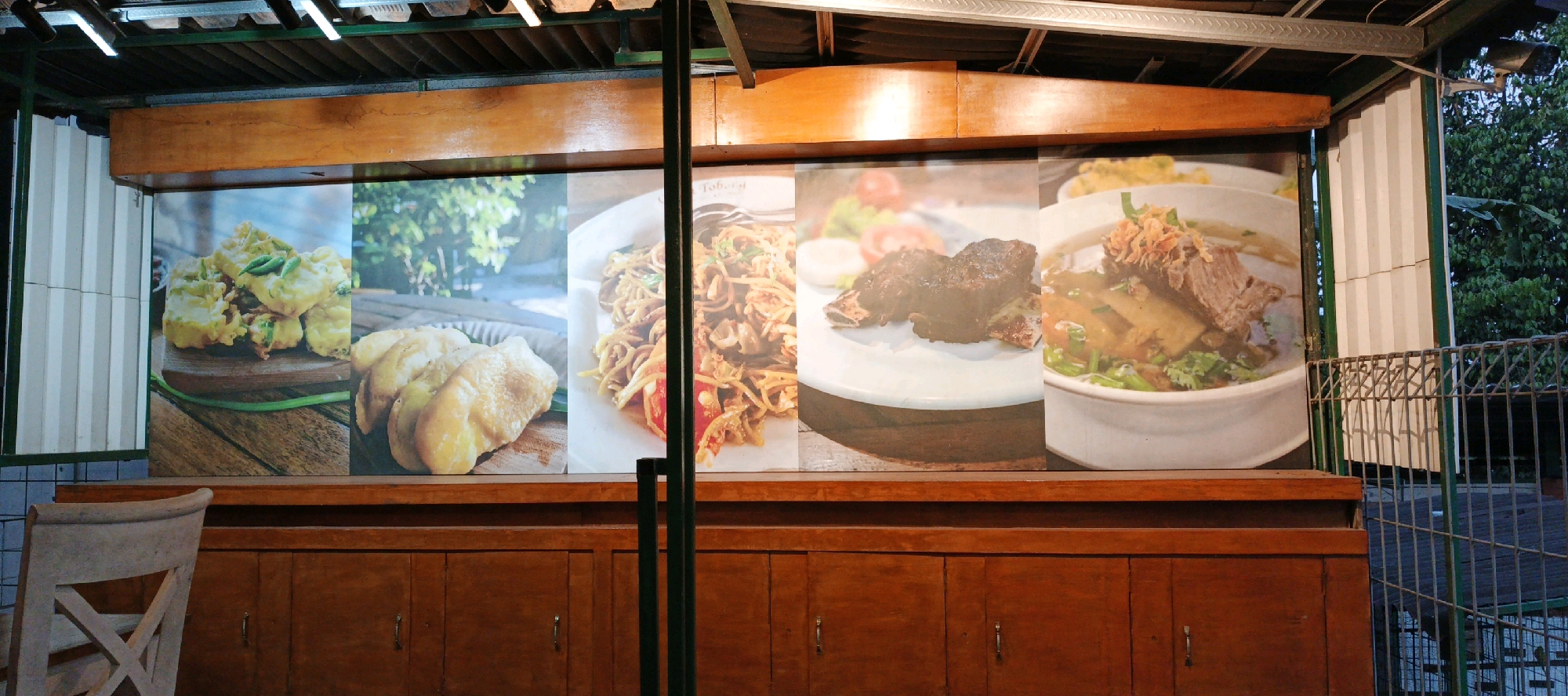 This is a medium shot, eye-level, outdoor photograph of a food display mural at what appears to be a restaurant or food stall. The mural, which spans across several panels, features close-up images of various Indonesian dishes. From left to right, the dishes depicted are: fried battered items (possibly tempeh or tofu) with green chilies, fried bananas, a plate of mie goreng (fried noodles), a plate of grilled ribs, and a bowl of soup with meat and vegetables. The mural is mounted on a wooden structure that runs along the top of a wooden cabinet with several drawers or doors. A light-colored wooden chair with a crisscross back is partially visible on the left side of the frame. In the background, a metal fence and lush green foliage suggest an outdoor setting, possibly a garden or a street scene. The overall lighting suggests it is either dusk or dawn, with artificial lights from above illuminating the scene. The specific location is Sidokarto, Indonesia.